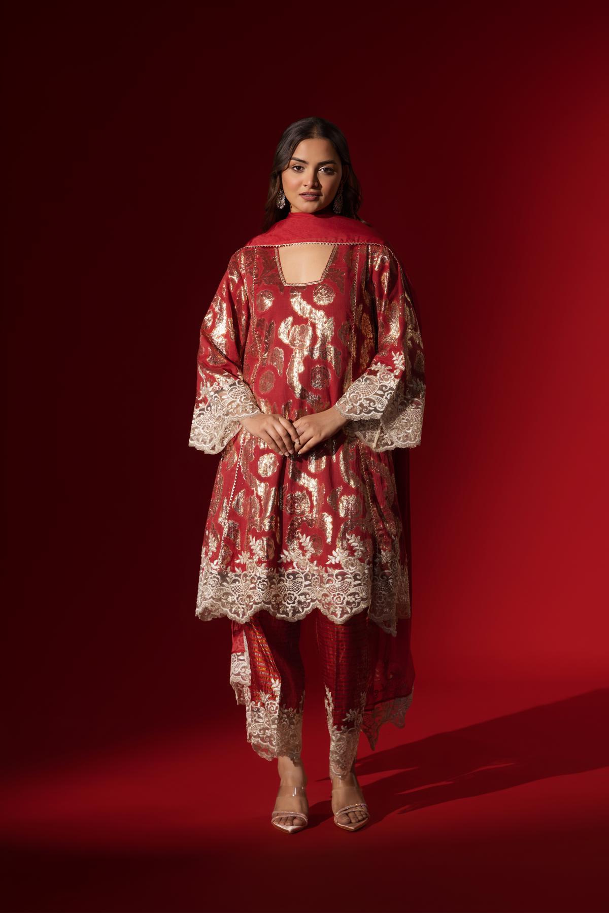 A woman poses in a maroon ethnic shimmer kurta set outfit with intricate white embroidery against a vibrant red backdrop.