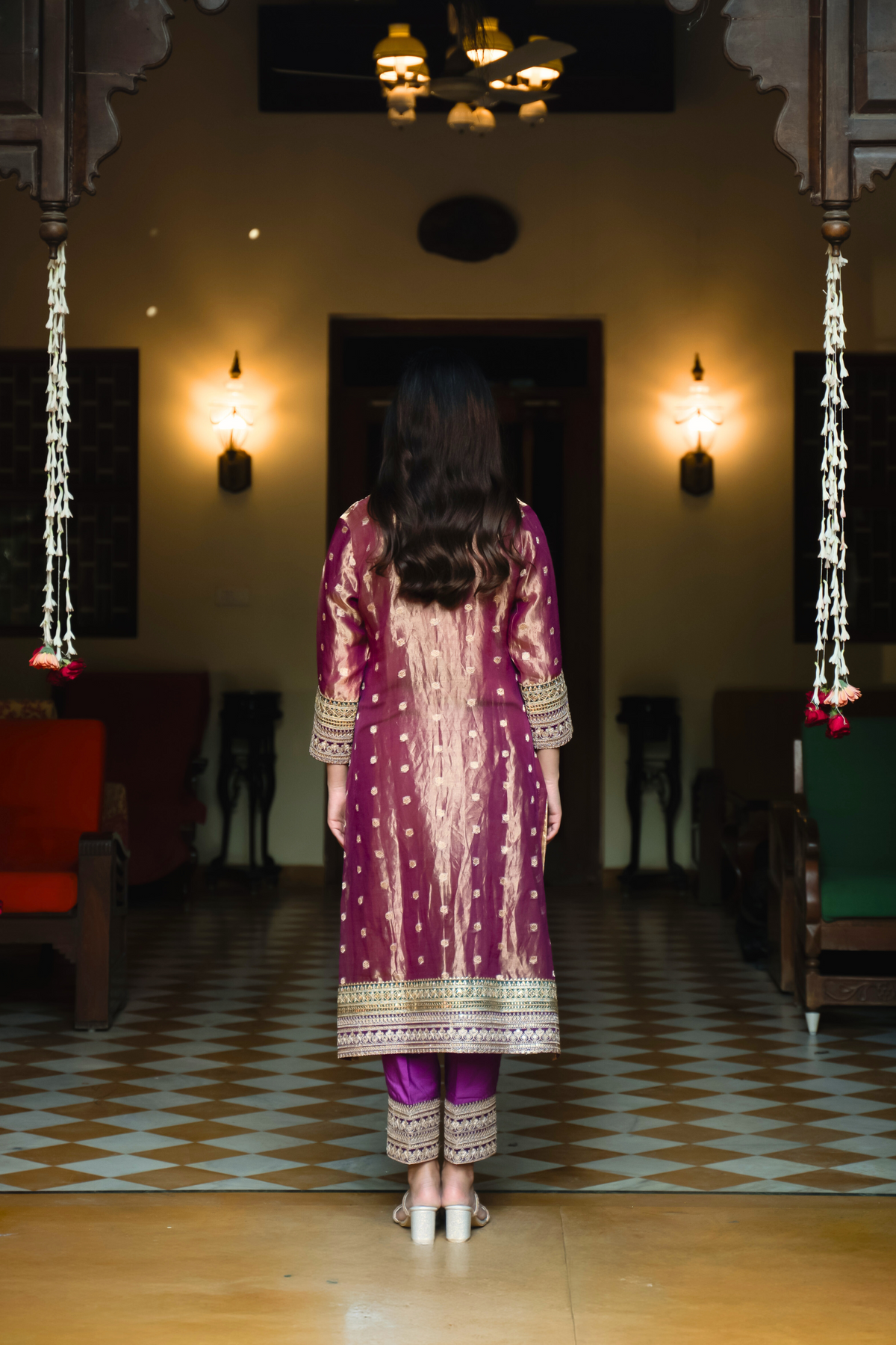 Back view of a woman in a traditional purple and gold anarkali outfit stands elegantly in a vintage-style room, surrounded by decor and plush seating.