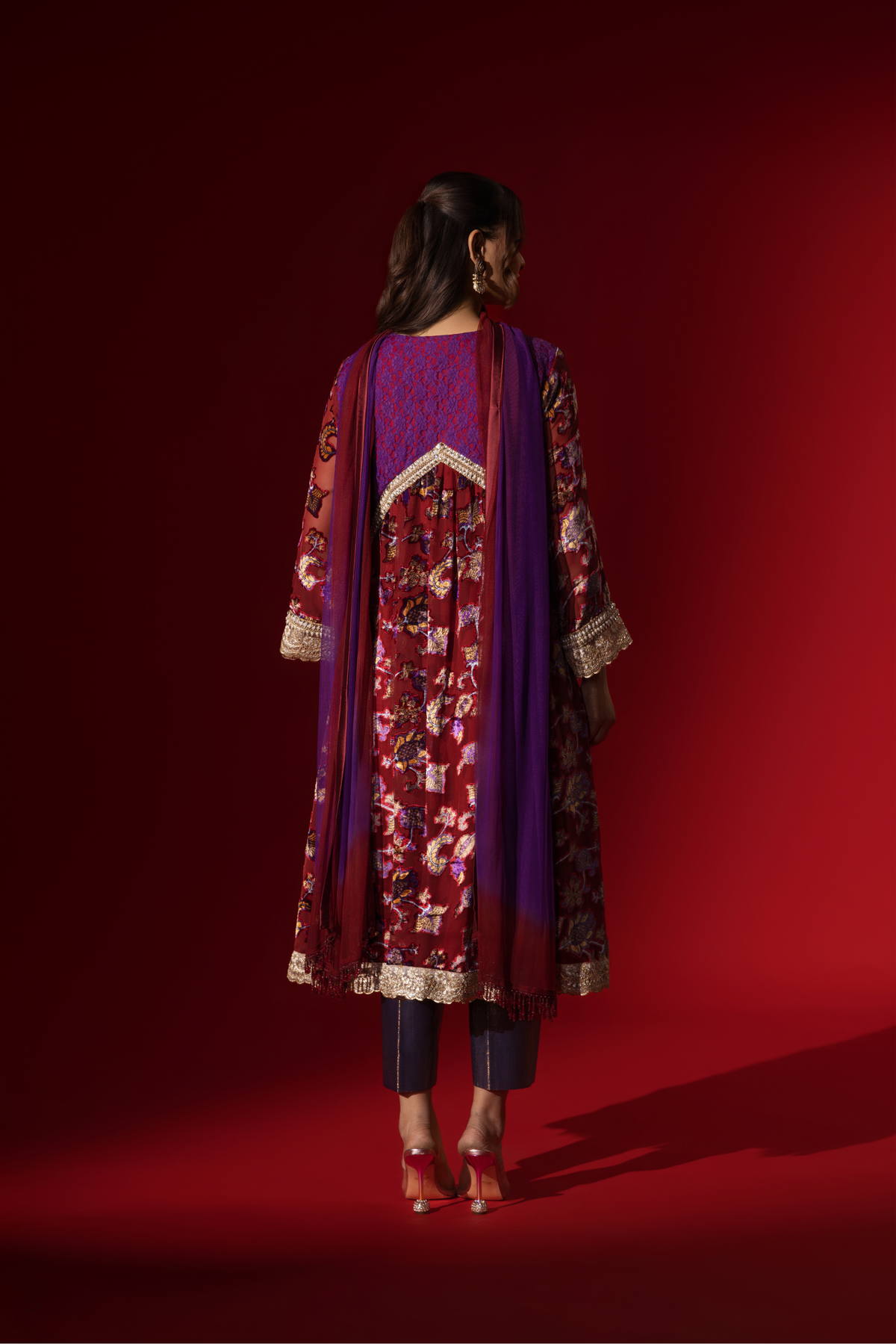 Back view of a model poses in a vibrant maroon floral velvet anarkali kurta set with purple accents and a golden trim, set against a rich red background.