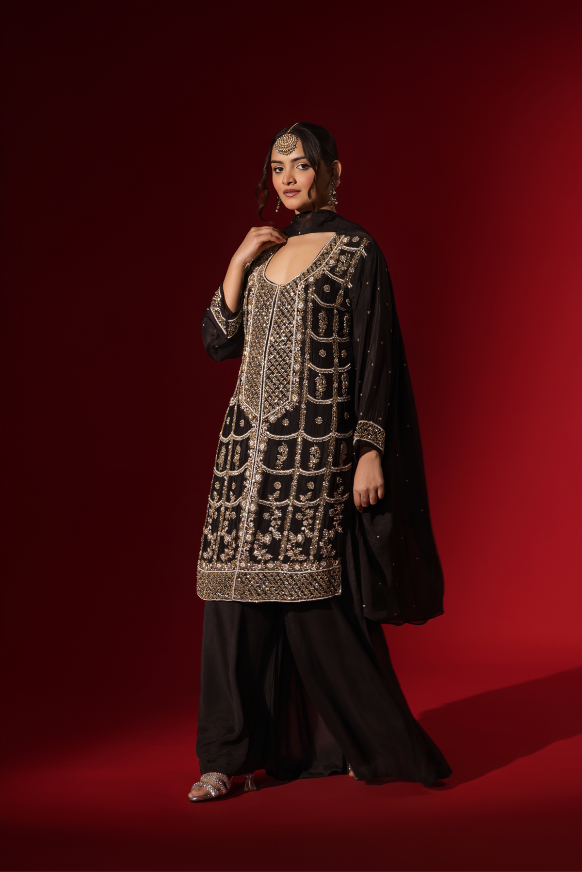 Side view of a woman showcases an elegant black embroidered kurta set with intricate gold embroidery against a rich red backdrop.