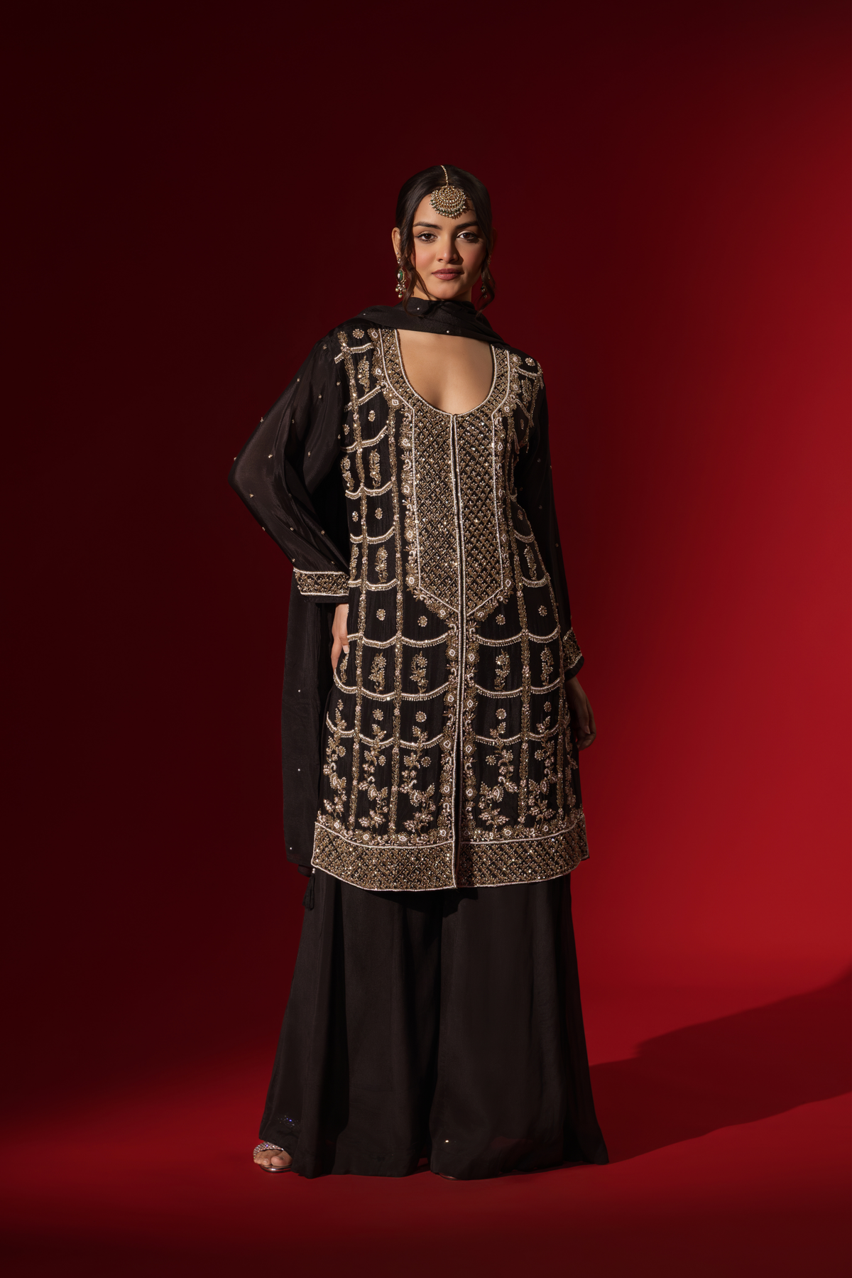 A woman showcases an elegant black embroidered kurta set with intricate gold embroidery against a rich red backdrop.