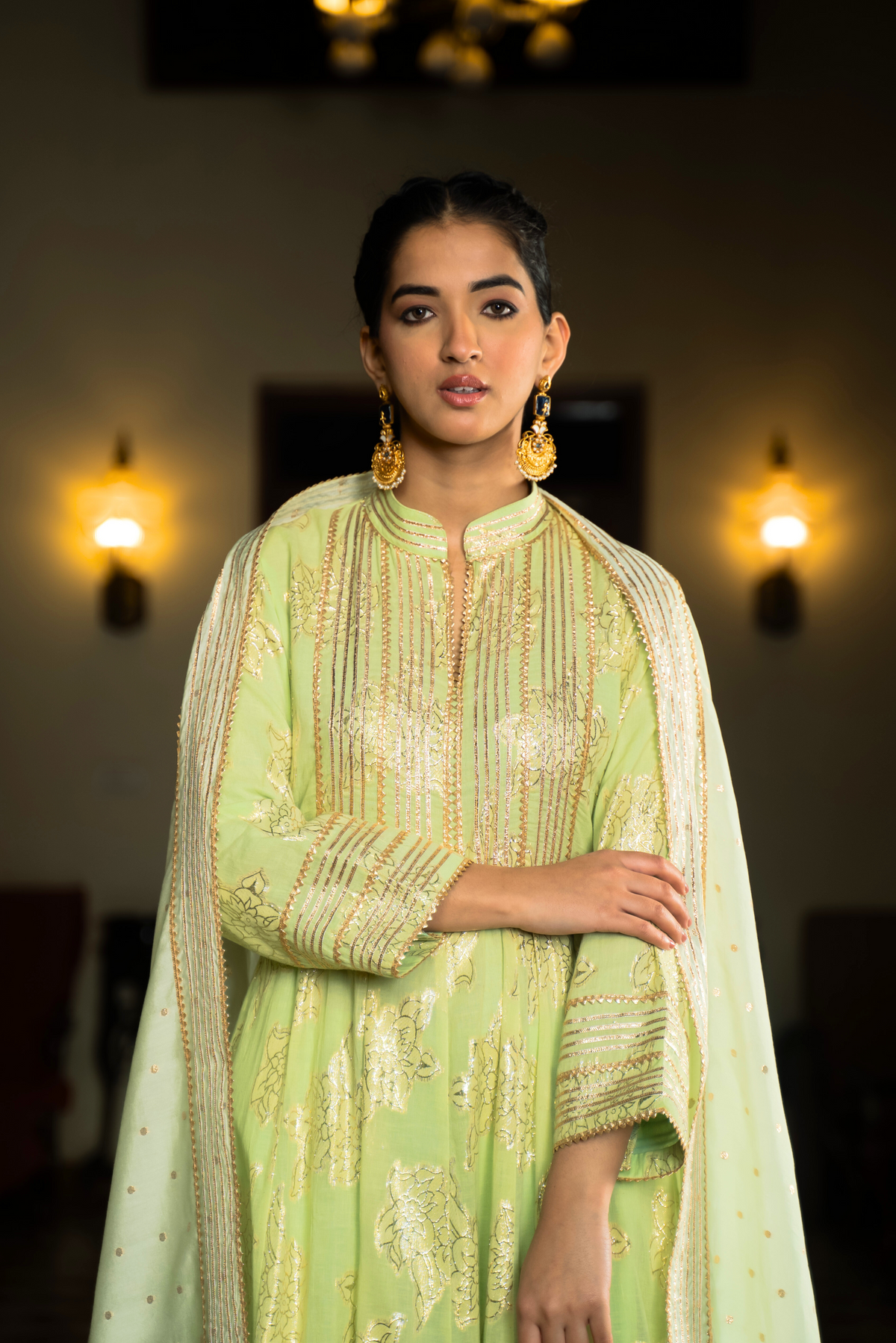 Closeup view of a woman in a mint green anarkali kurta set outfit stands elegantly in a beautifully decorated indoor space with warm lighting and classic furnishings.