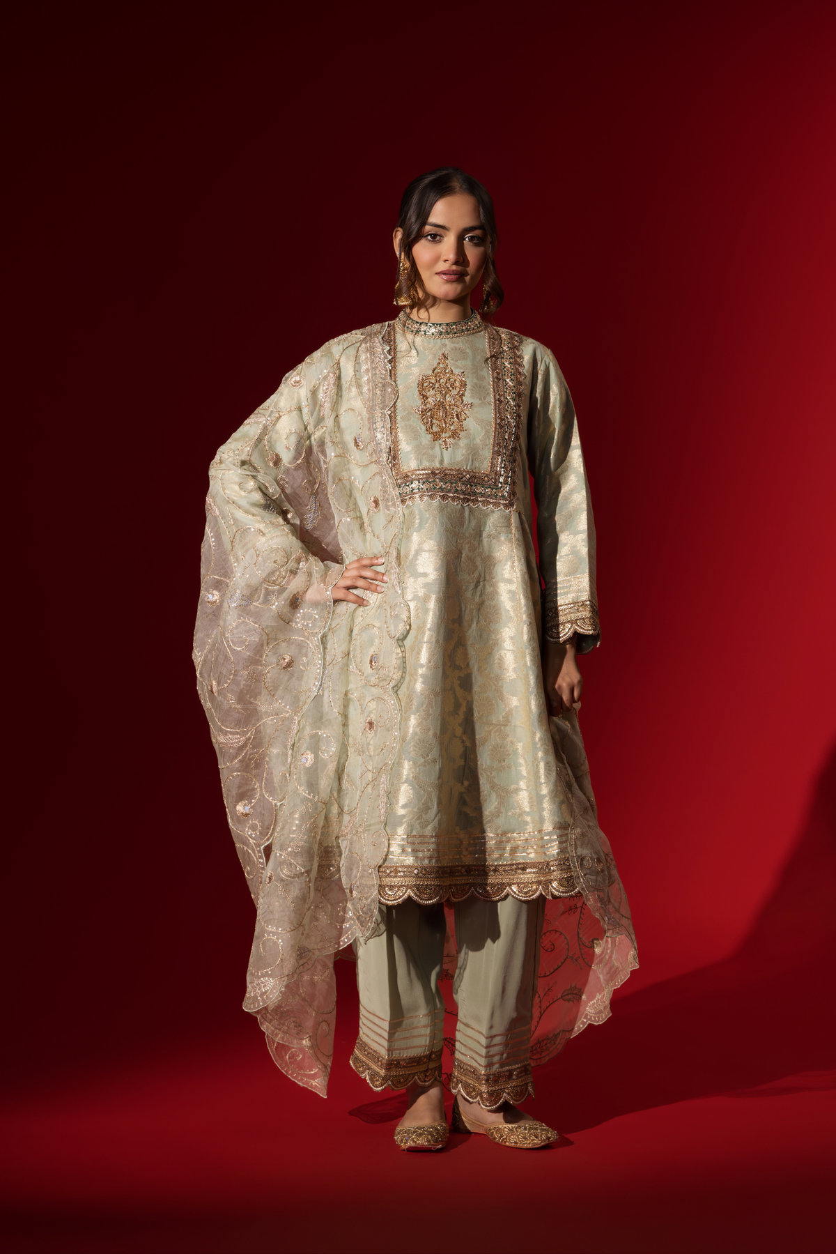 A woman wearing an elegant mint green embroidered sequin brocade kurta set with gold accents and a sheer dupatta, set against a rich red background