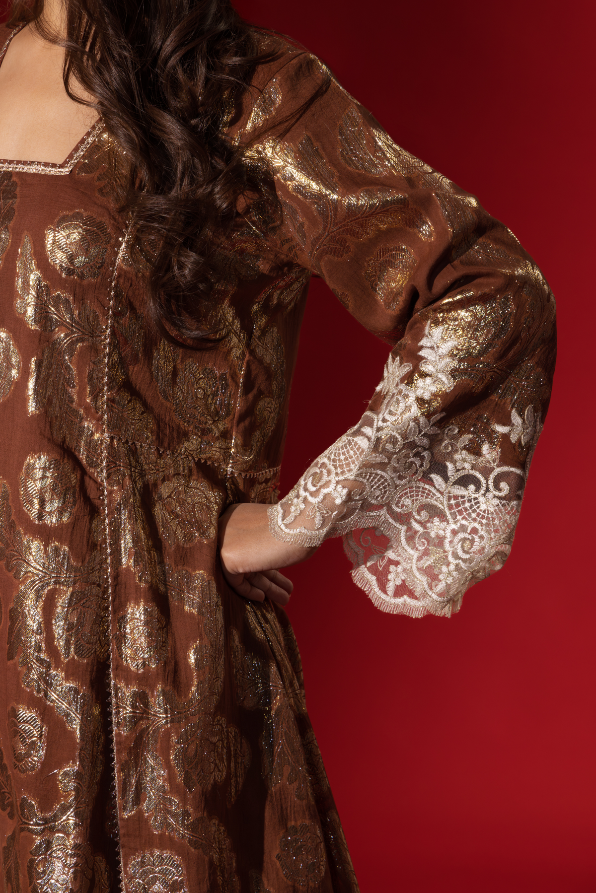 Closeup view of a women's hand in a brown ethnic shimmer kurta set outfit with intricate white embroidery against a vibrant red backdrop.