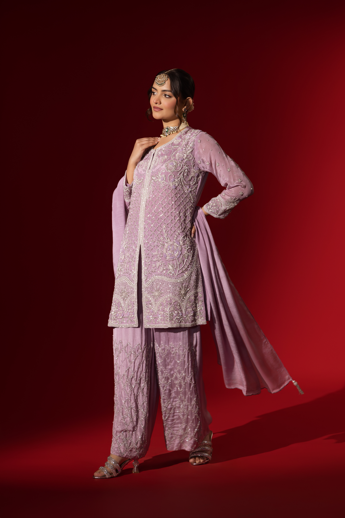 Side view of a woman stands confidently in a beautifully lavender embroidered kurta set, against a rich red background.