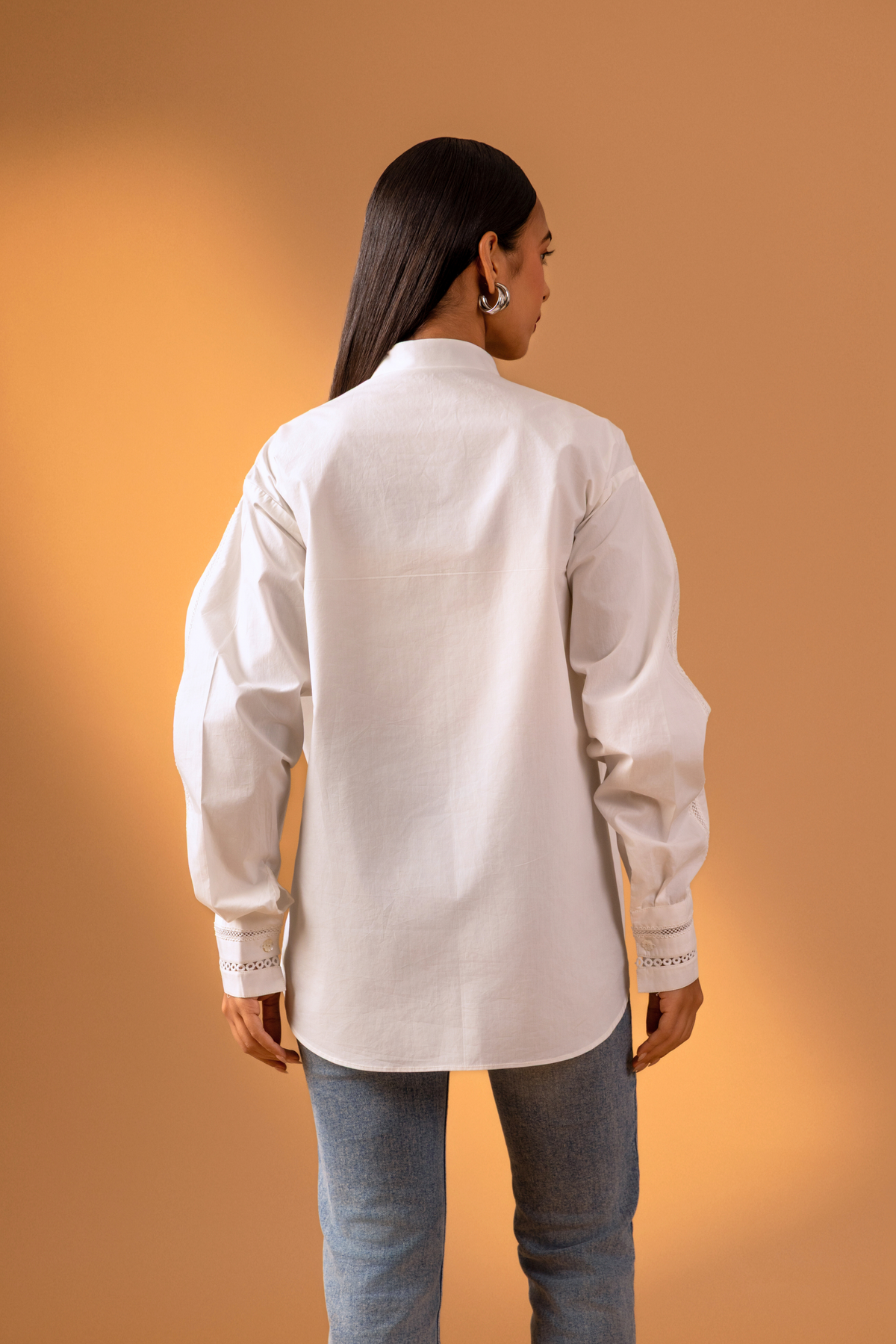 Back view women stands confidently in a stylish white button-up shirt with intricate details and puffed sleeves, paired with blue jeans.