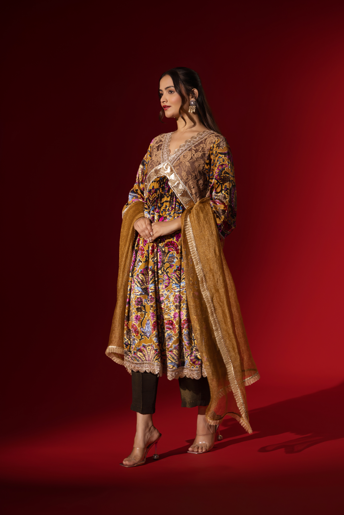 Side view of a woman stands confidently in a colorful, embellished regal printed velvet kurta set with a flowing golden dupatta against a vibrant red backdrop.