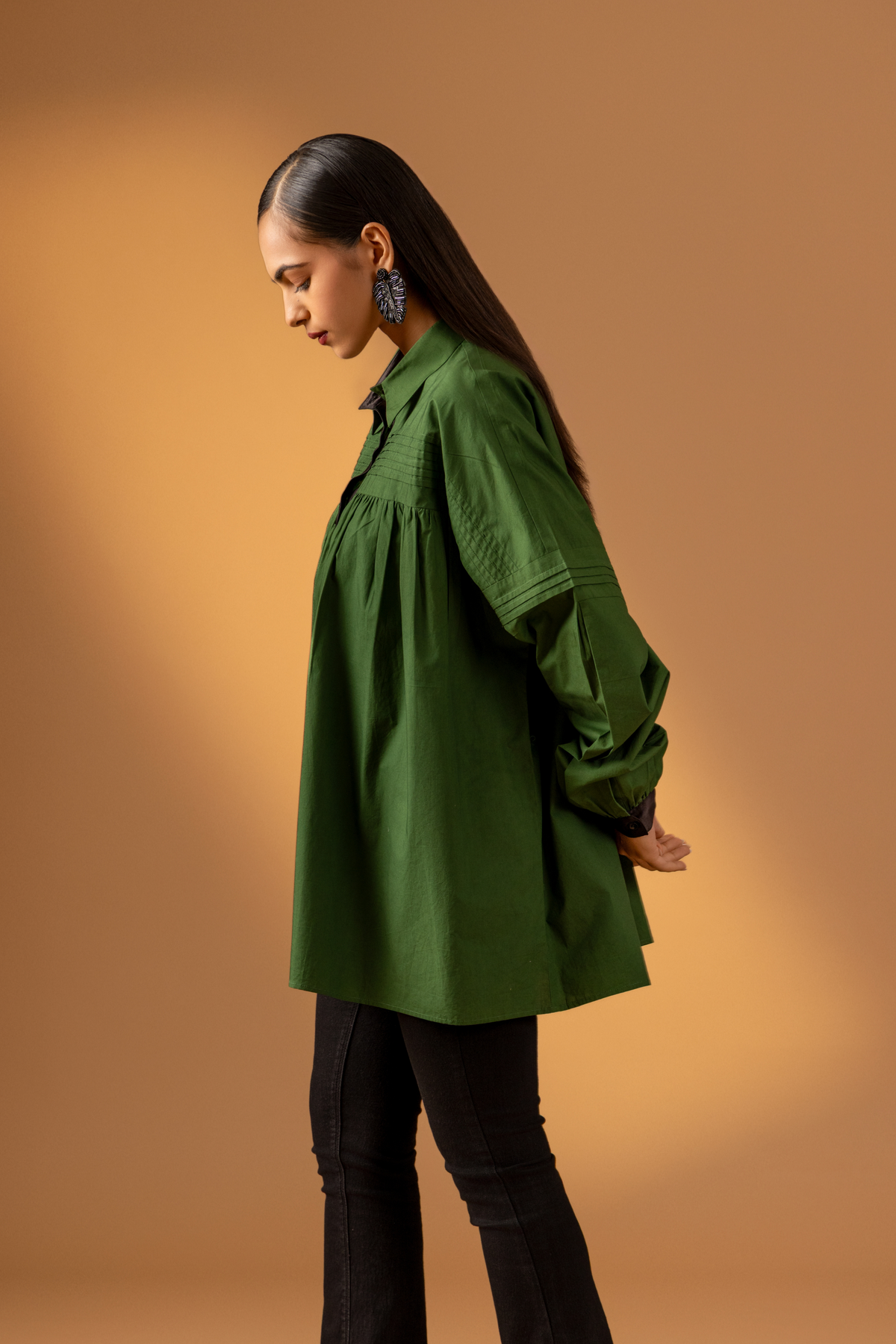 Side view of a woman stands against a warm beige background, wearing a loose green shirt with a black button front and black flared pants .