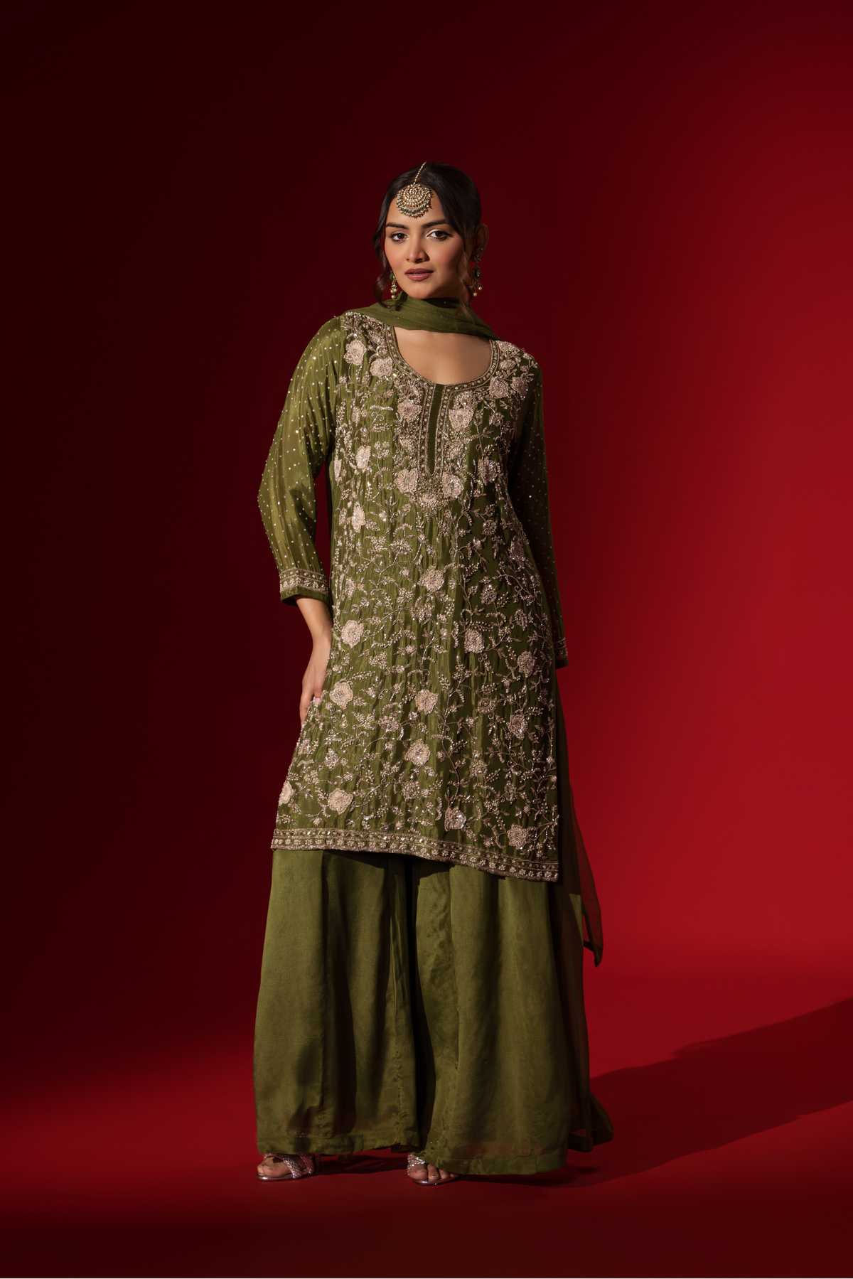 A woman poses in an elegant olive green embroidered kurta set with intricate floral embroidery, set against a rich red backdrop.