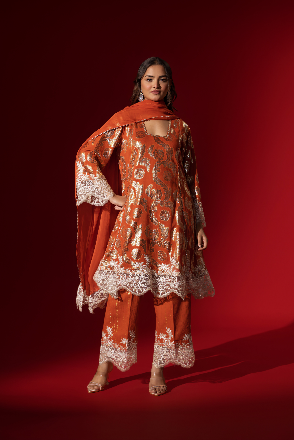 A woman poses in a orange ethnic shimmer kurta set outfit with intricate white embroidery against a vibrant red backdrop.
