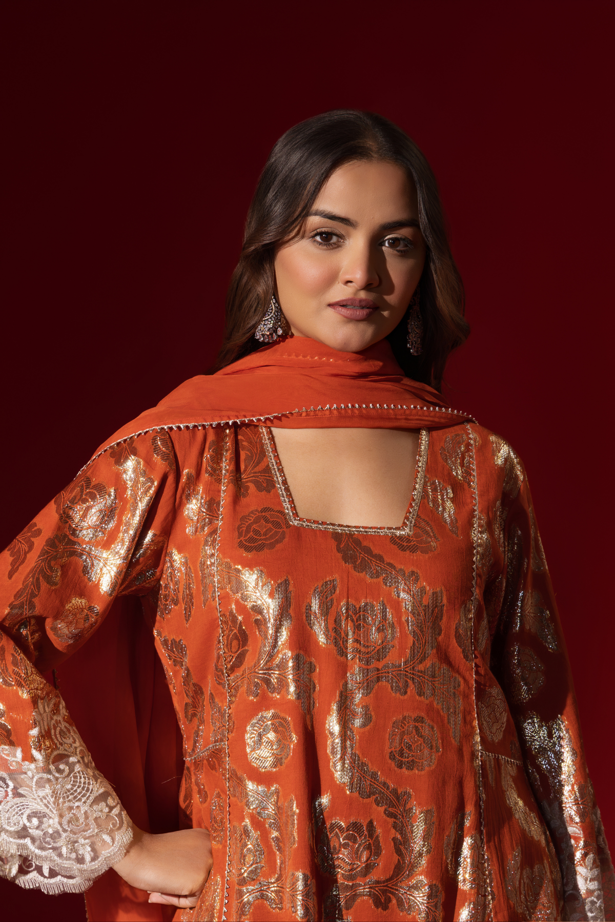 Closeup view of a woman poses in a orange ethnic shimmer kurta set outfit with intricate white embroidery against a vibrant red backdrop.