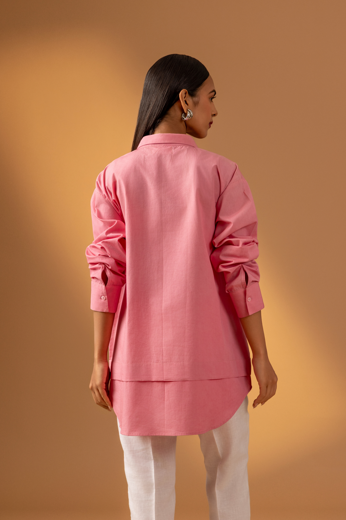 Back view of a woman wearing a pink shirt paired with white trousers, and sandals against a warm, neutral background.