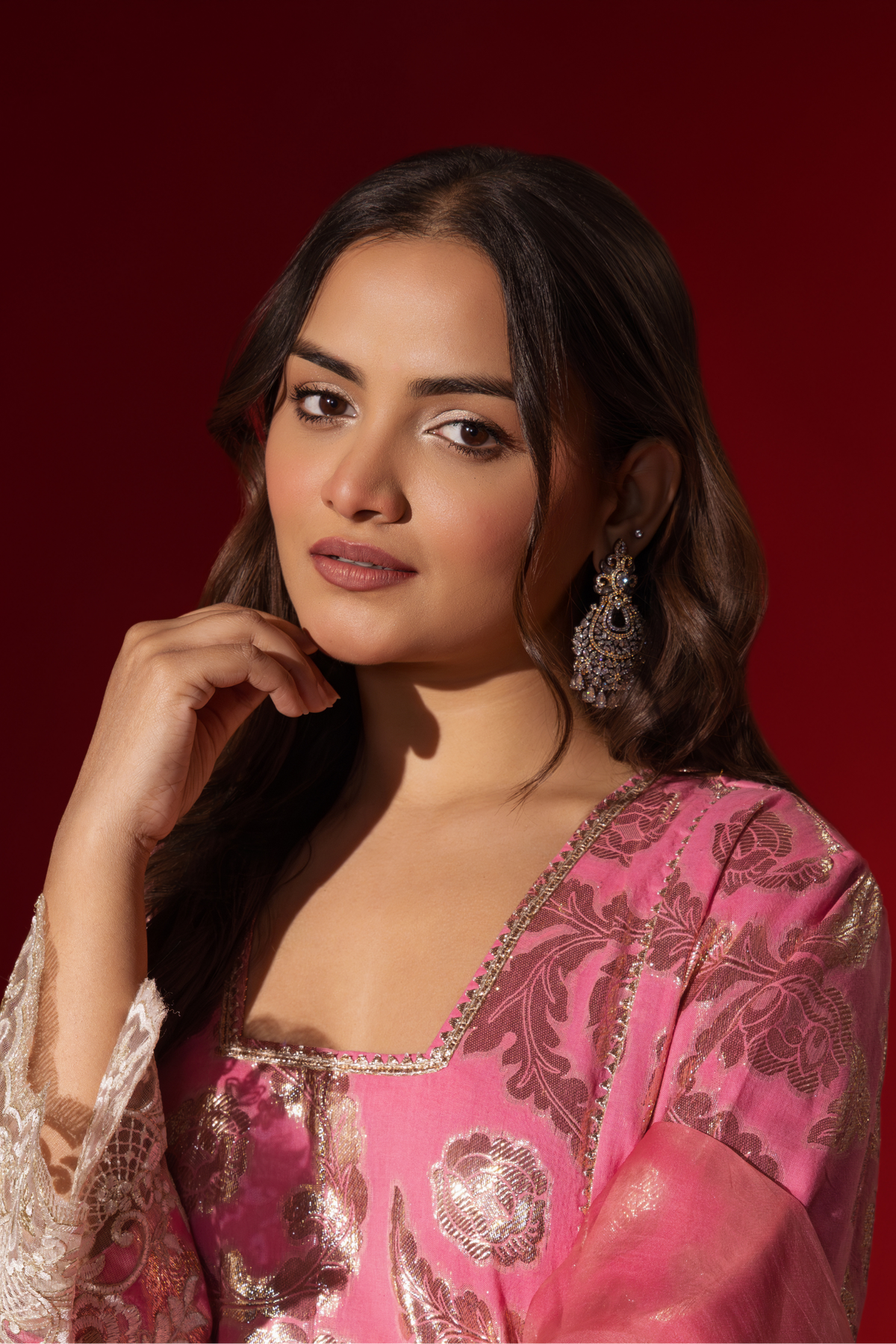 Closeup view of a woman poses in a pink ethnic shimmer kurta set outfit with intricate white embroidery against a vibrant red backdrop.