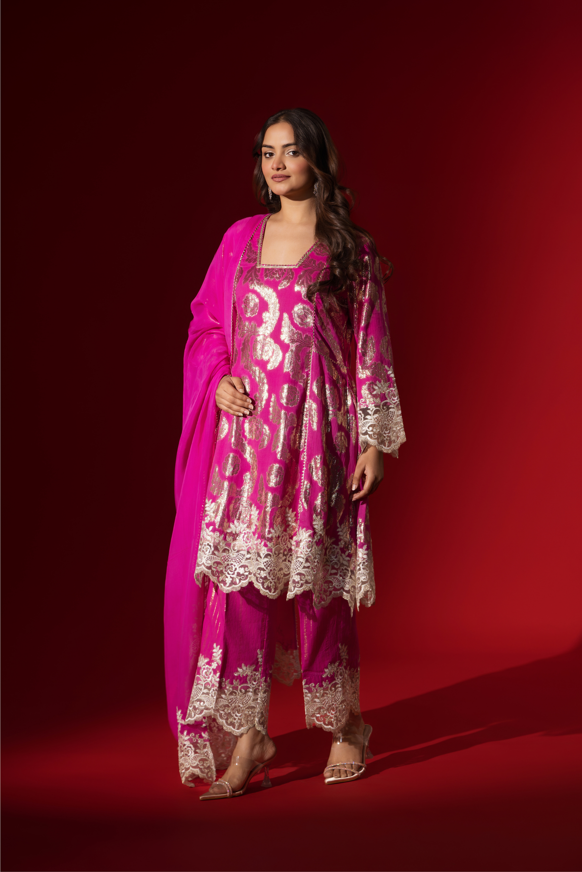 Side view of a woman in an elegant pink shimmer kurta set with intricate golden embroidery stands against a vibrant red background.