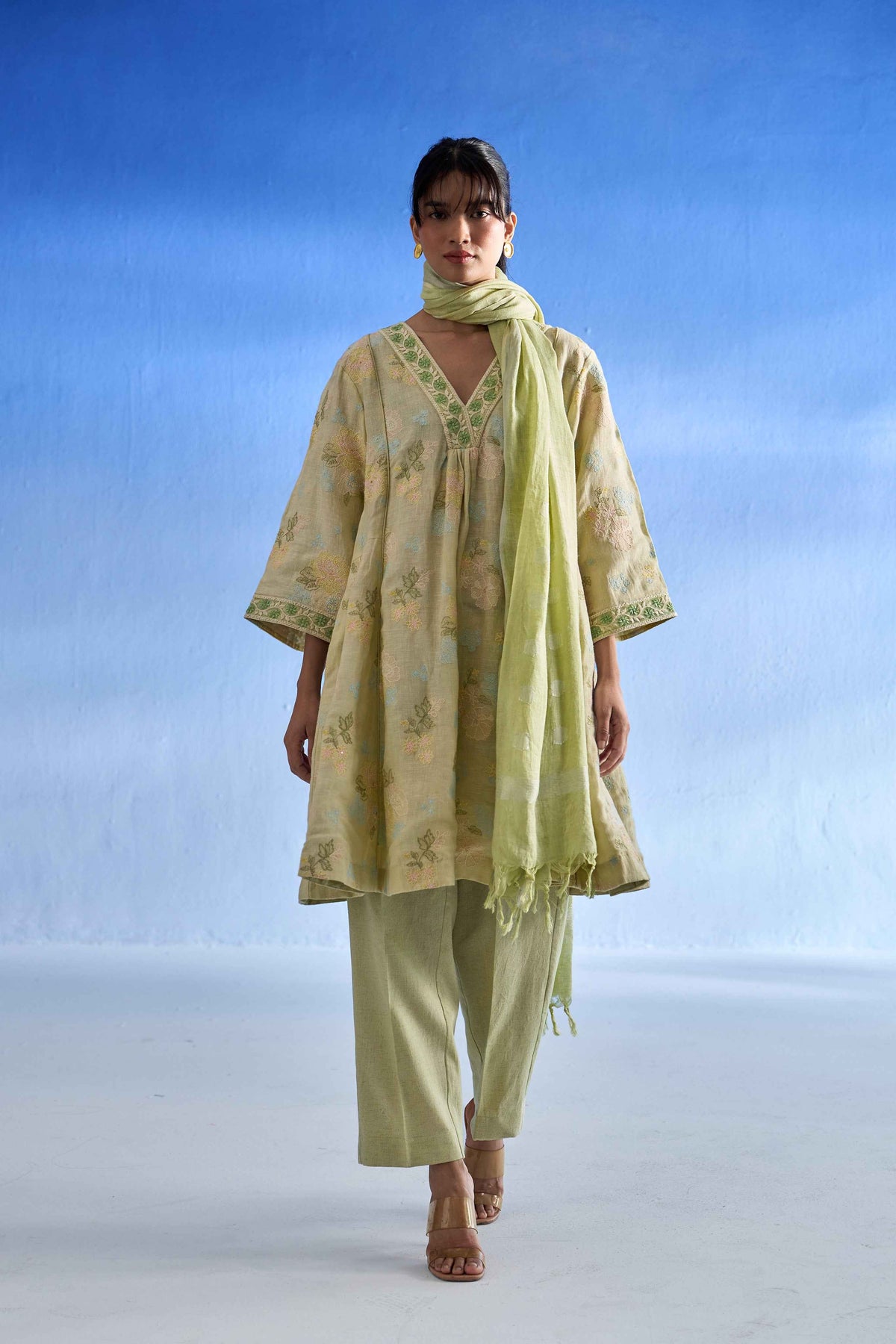 A woman modeling a pistachio green and beige floral printed kurta set with matching pants and scarf against a blue gradient studio background.