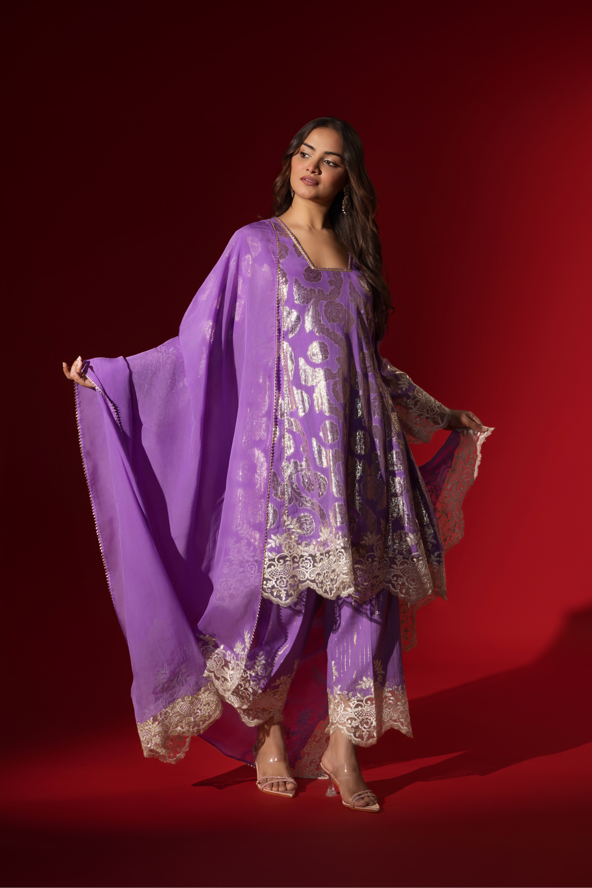 Side view of a woman poses in a purple ethnic shimmer kurta set outfit with intricate white embroidery against a vibrant red backdrop.