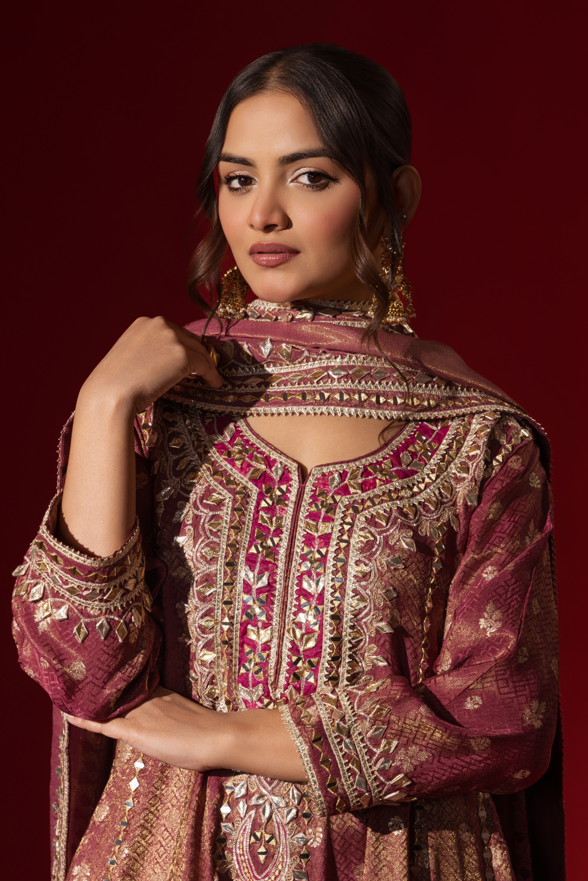 Closeup view of a woman stands elegantly in a rich maroon embroidered brocade kurta set adorned with intricate gold embroidery, against a deep red background.