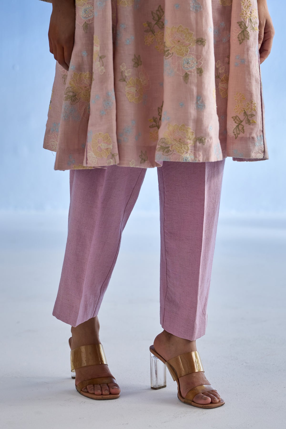 A person wearing a rose bloom floral printed linen kurta set, dusty purple straight-leg trousers, and gold heeled sandals against a pale blue studio background.