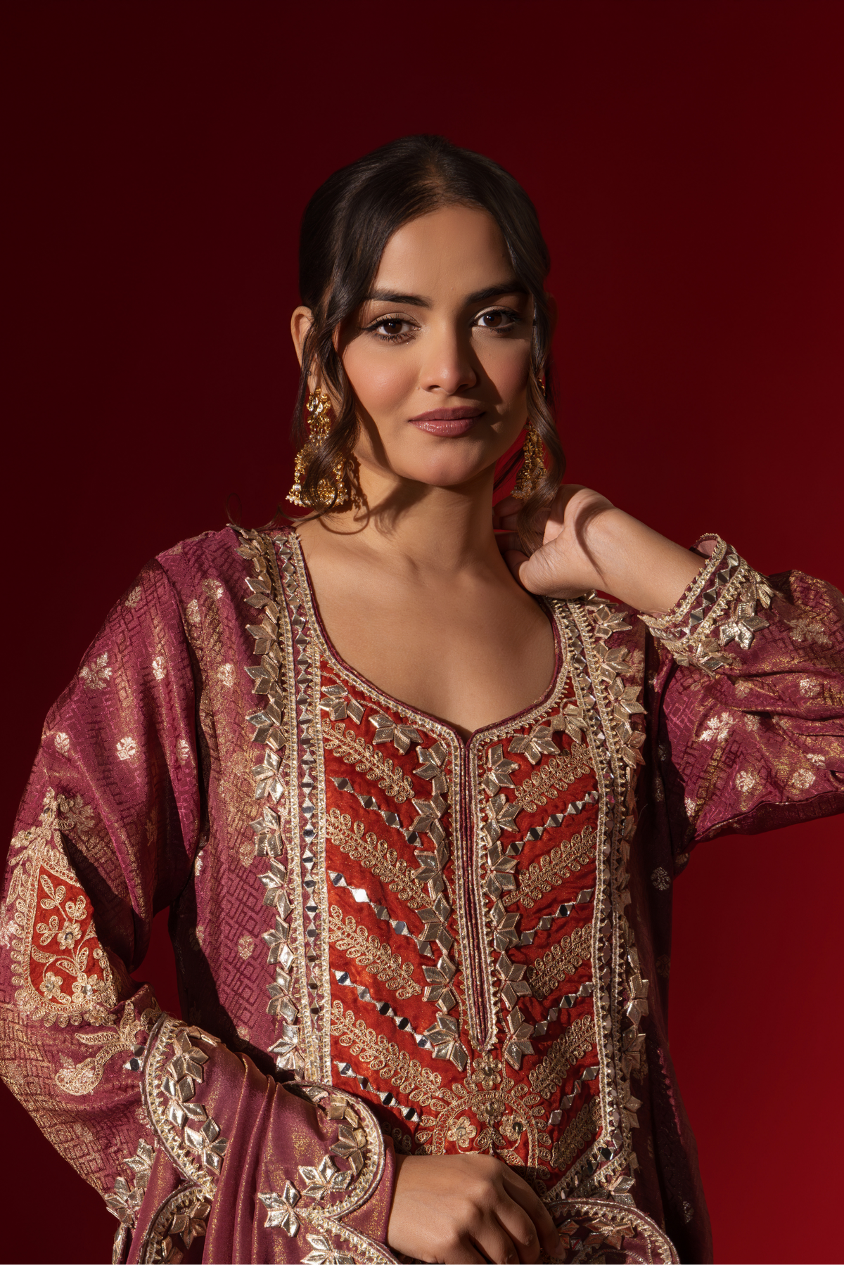 Closeup view of a woman wearing a richly embellished maroon embroidered brocade kurta set outfit with intricate gold detailing against a striking red background.