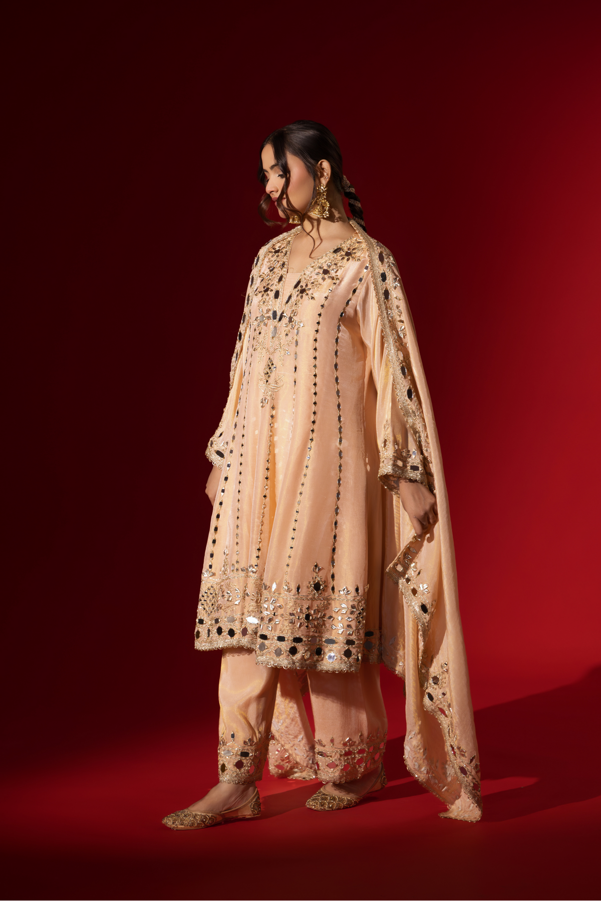 Side view of a woman stands elegantly in a beige embroidered kurta set with intricate patterns, complemented by a matching dupatta against a bold red background.