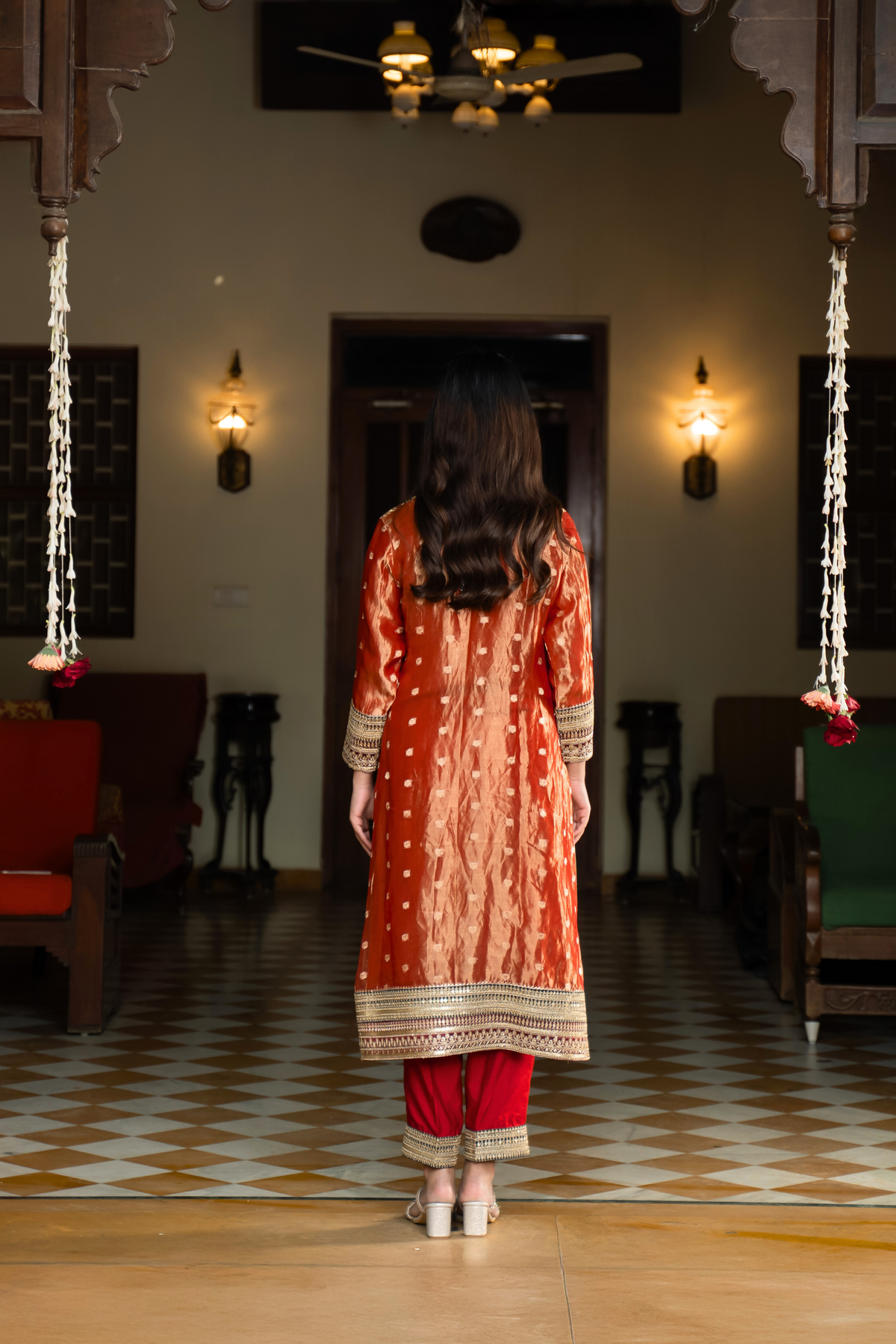 Back view of a person in a traditional orange and golden banarsi silk kurta set outfit stands elegantly in a beautifully decorated room with vintage furnishings.