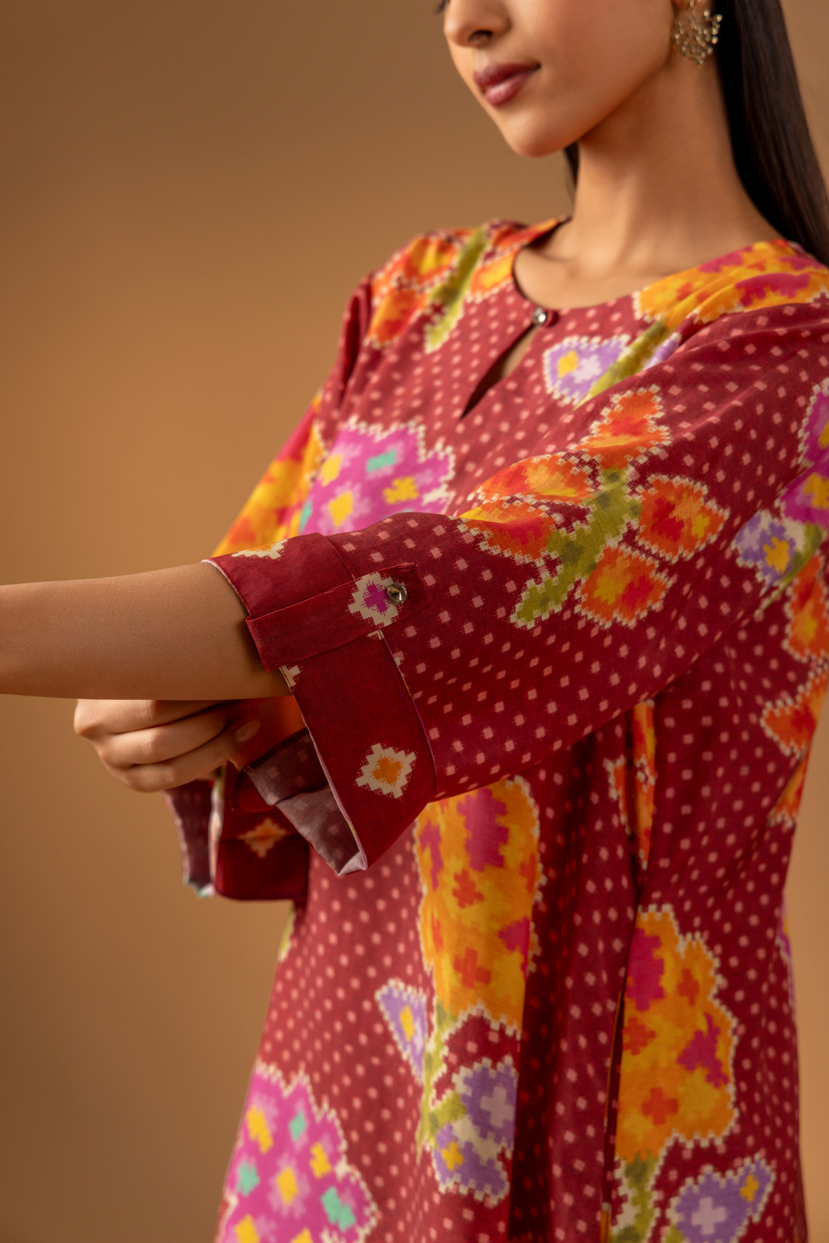 Closeup view of a model wears a vibrant floral co-ord set over matching patterned pants, and showing her hand set against a warm, neutral backdrop.