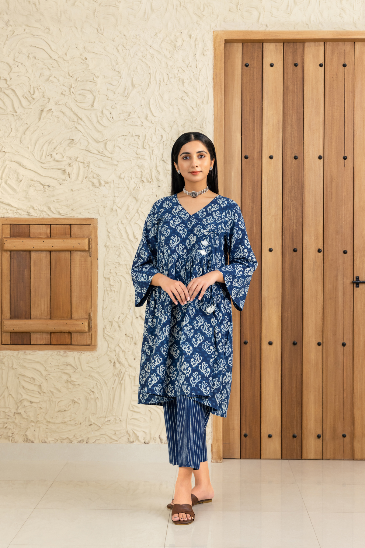 Woman wearing a blue and white printed co-ord set with a V-neck and tassel details, standing against a wooden and textured beige background.