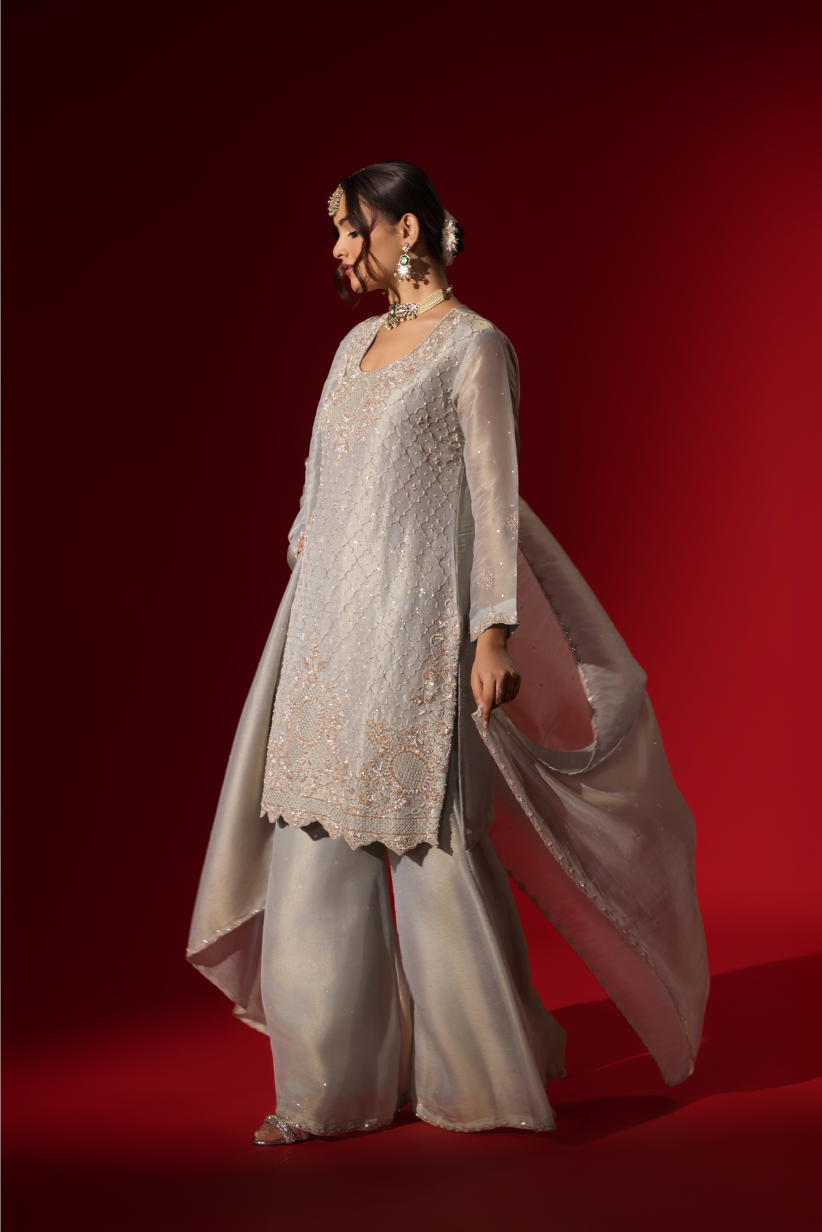 Side view of a woman stands elegantly in a shimmering silver grey embroidered gharara set, featuring intricate embroidery, paired with matching wide-legged trousers and a flowing shawl.