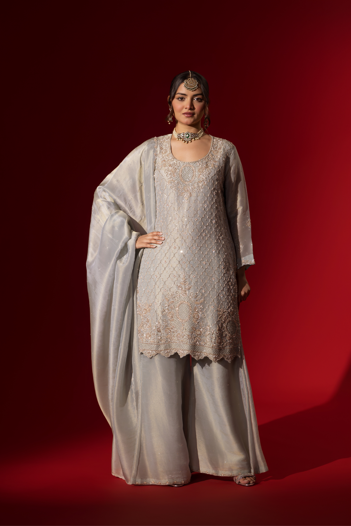 A woman stands elegantly in a shimmering silver grey embroidered gharara set, featuring intricate embroidery, paired with matching wide-legged trousers and a flowing shawl.