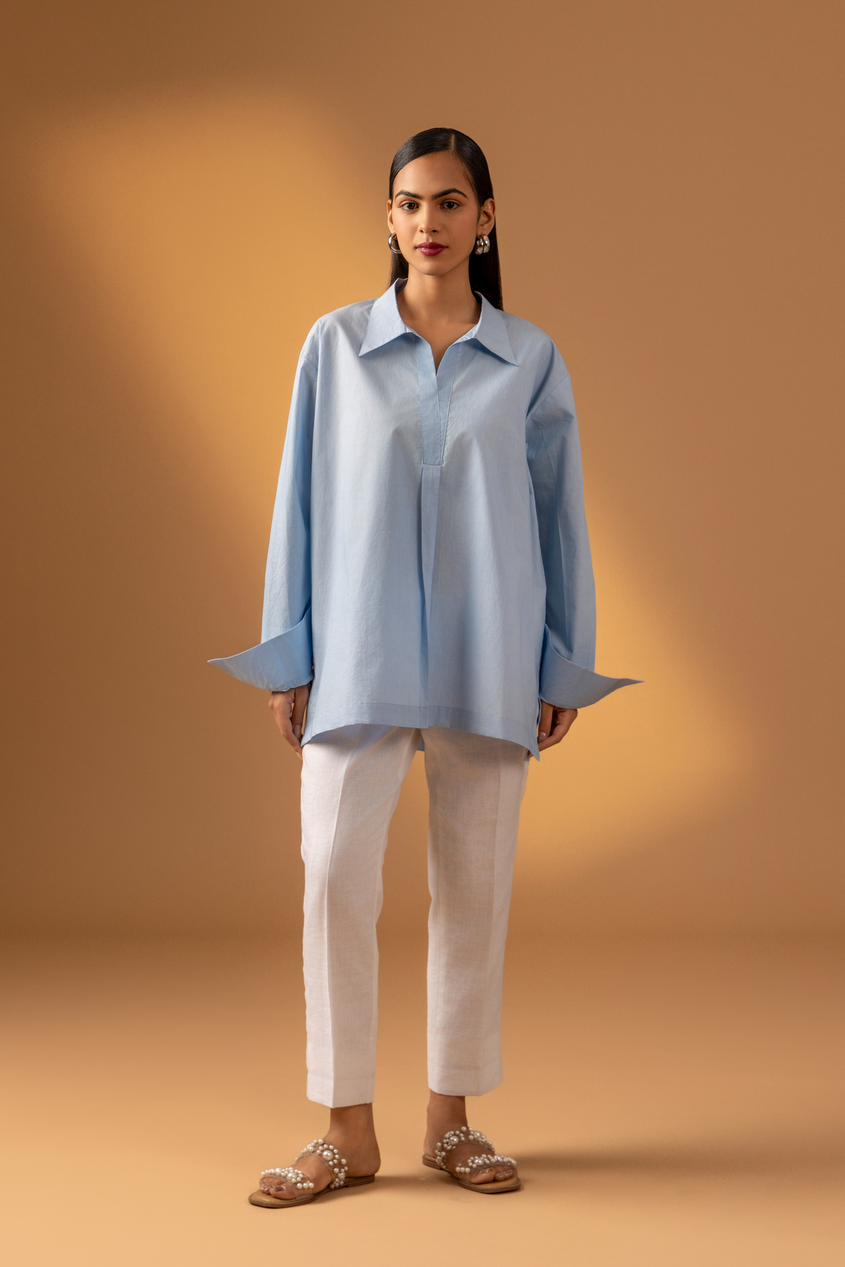 A women stands confidently wearing a light blue oversized shirt, white pants, and stylish sandals, against a warm beige background.