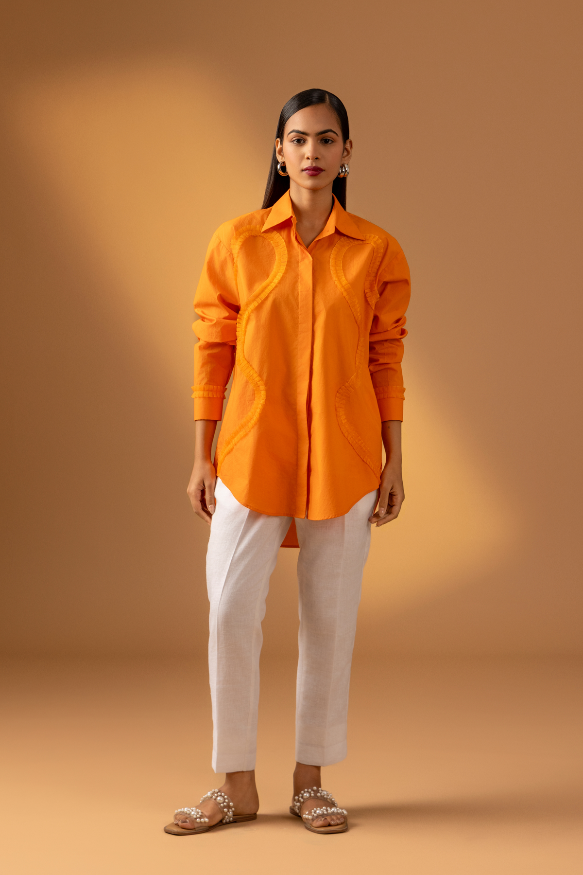 A Woman stands confidently in an orange shirt paired with white trousers, and sandals against a warm, neutral background.