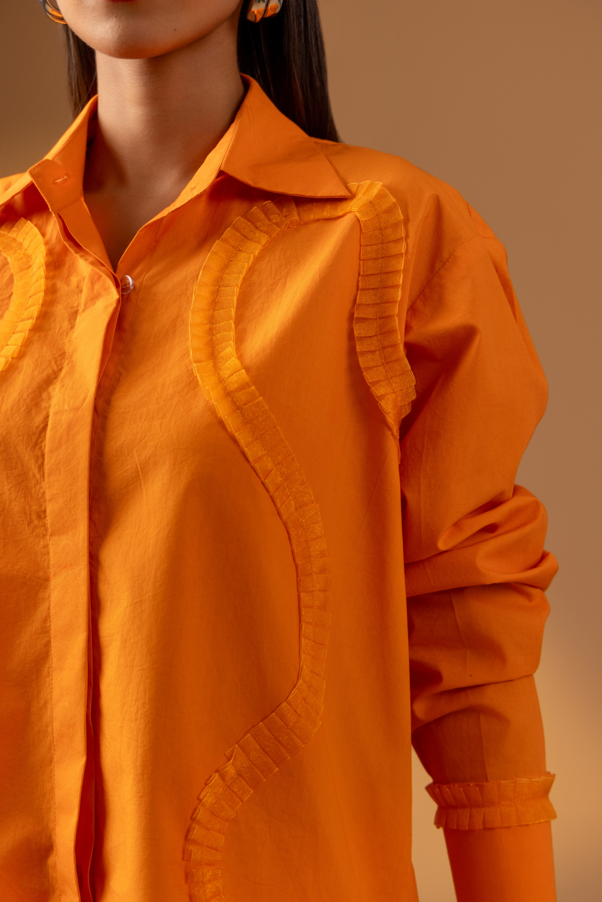 Closeup view of a woman stands confidently in an orange shirt paired with white trousers, and sandals, against a warm, neutral background.
