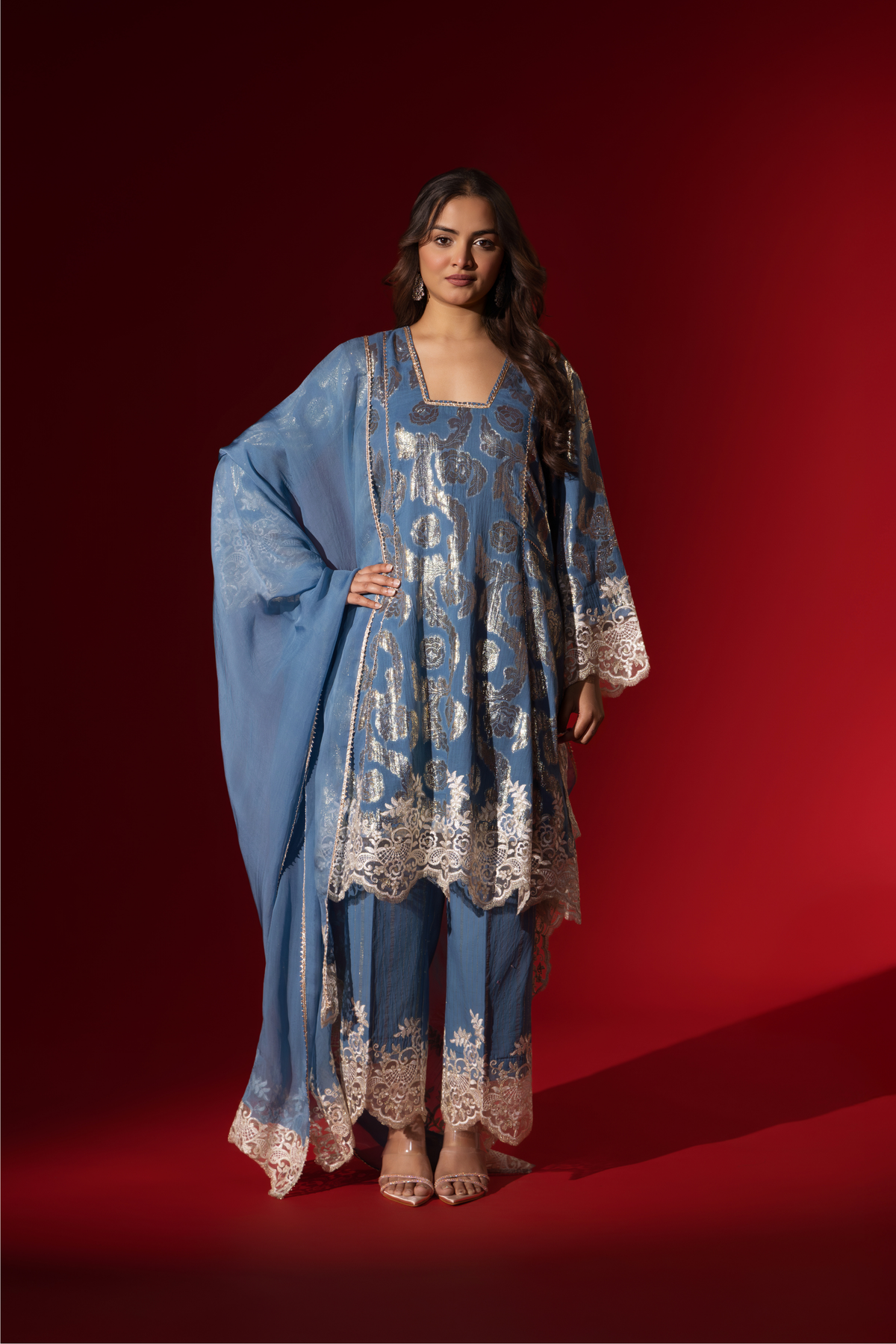 A woman poses in a blue ethnic shimmer kurta set outfit with intricate white embroidery against a vibrant red backdrop.