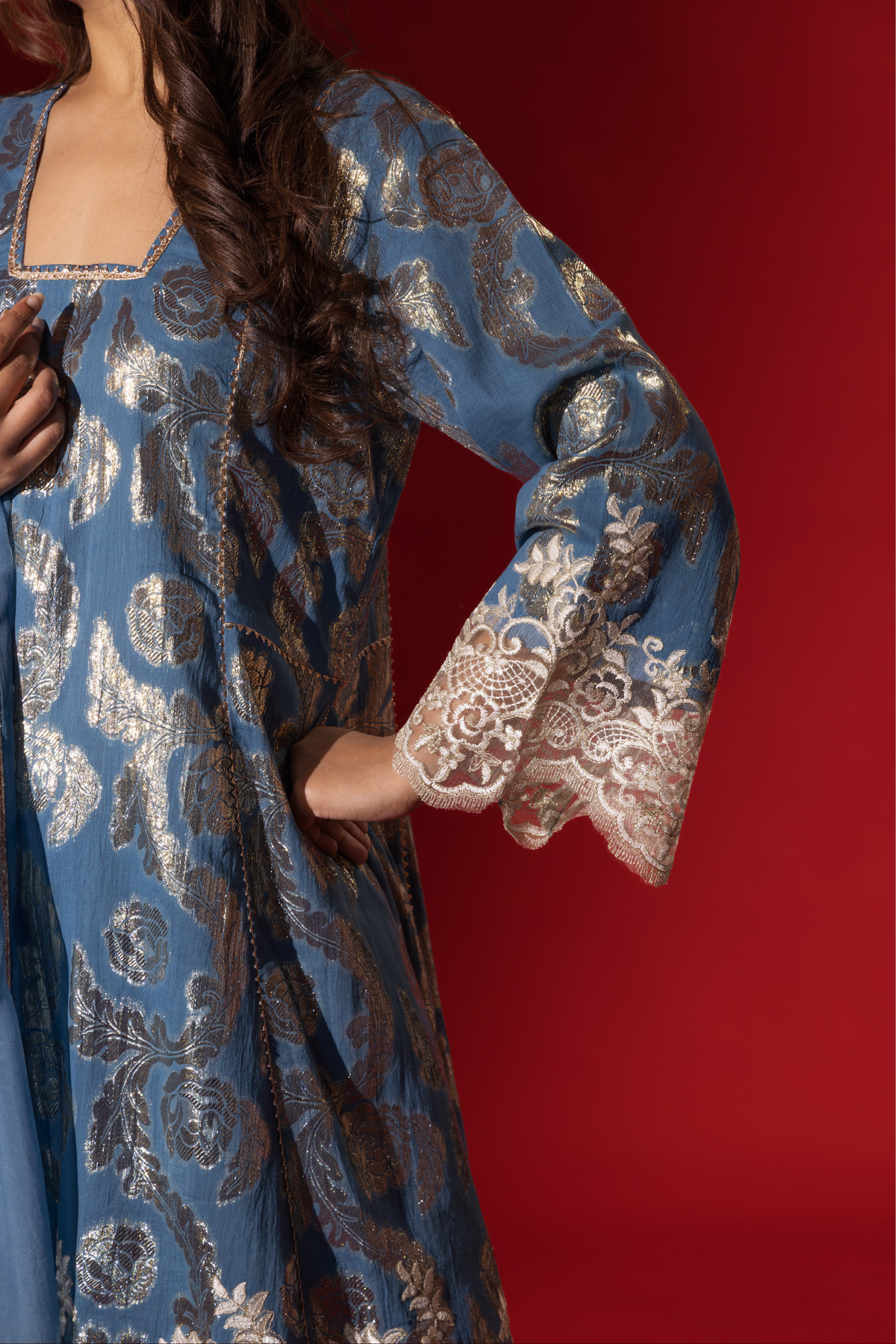Closeup view of a woman's hand in a blue ethnic shimmer kurta set outfit with intricate white embroidery against a vibrant red backdrop.