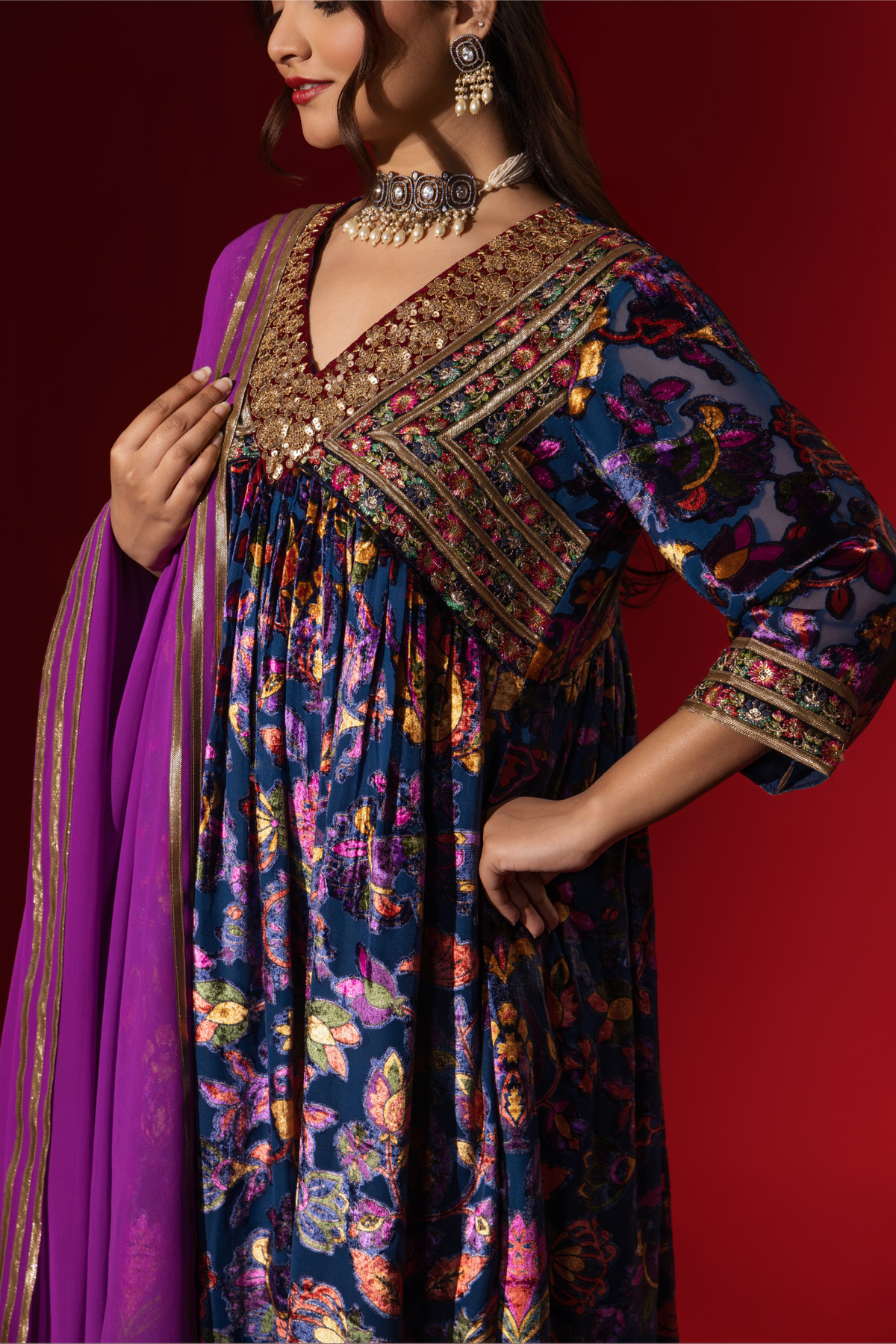 Closeup view of a woman's hand in a vibrant, embroidered purple printed velvet anarkali kurta set, paired with a matching dupatta against a rich red backdrop.