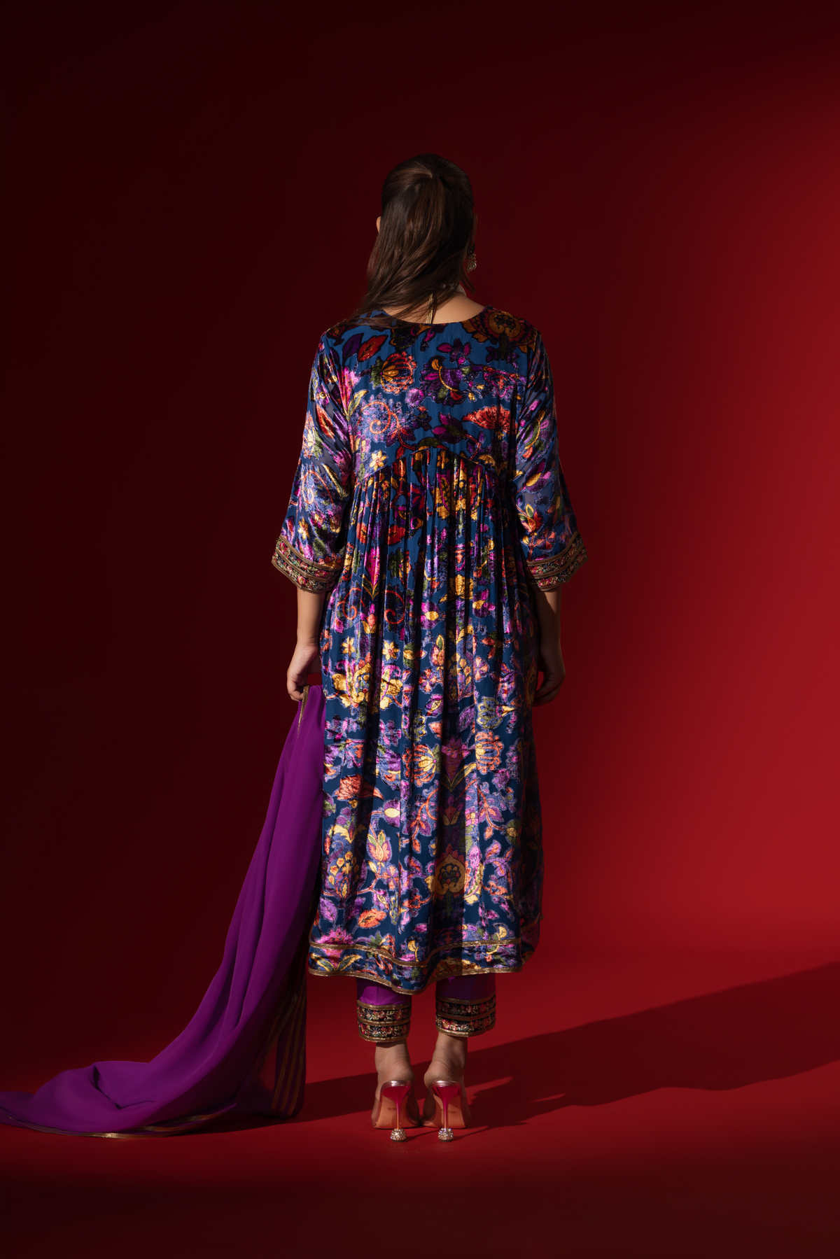 Back view of a woman stands confidently in a vibrant, embroidered purple printed velvet anarkali kurta set, paired with a matching dupatta against a rich red backdrop.