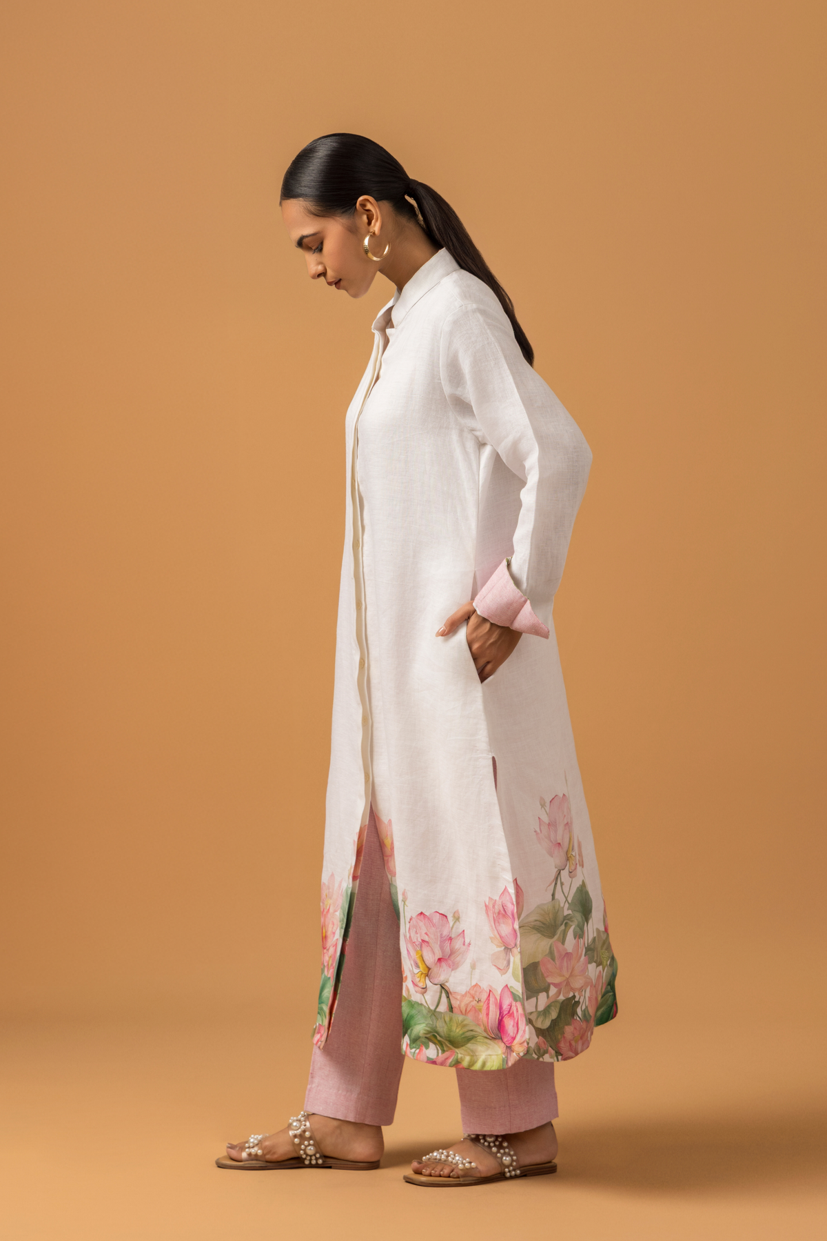 Side view of a model wearing a white lotus print linen co-ord set with floral patterns at the hem, paired with pink trousers and standing against a warm orange backdrop.