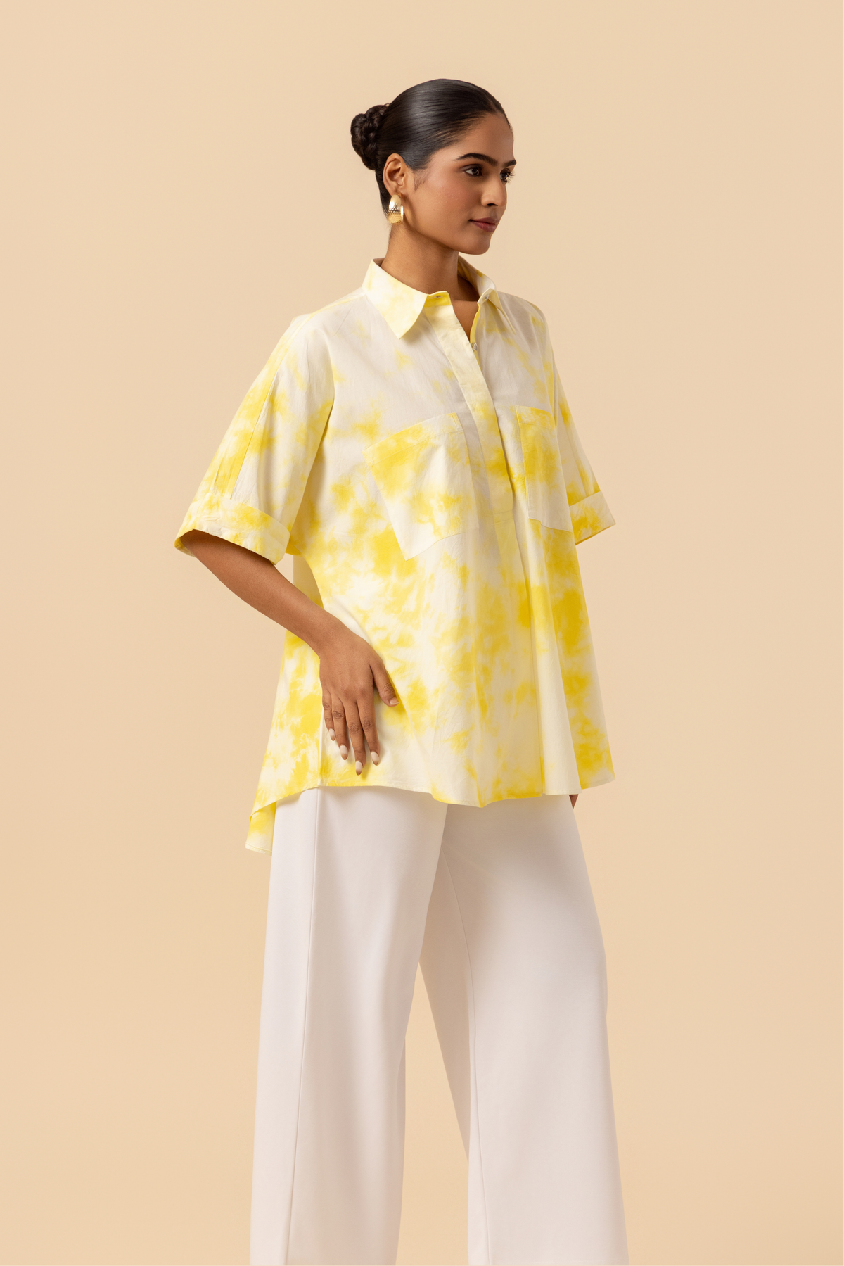 Right side view of a model poses confidently in a yellow tie-dye oversized shirt and white wide-leg pants against a soft beige background.