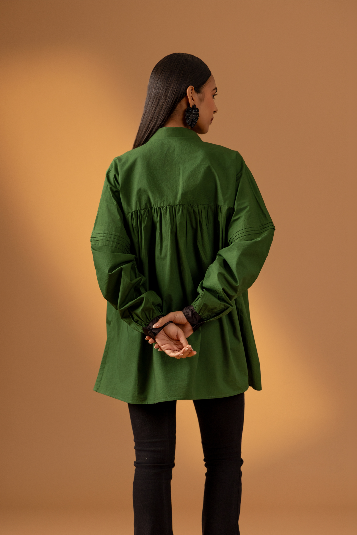 Back view of a woman stands against a warm beige background, wearing a loose green shirt with a black button front and black flared pants .