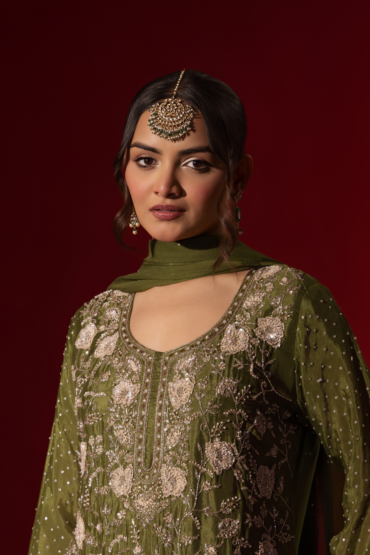 Closeup view of a woman poses in an elegant olive green embroidered kurta set with intricate floral embroidery, set against a rich red backdrop.