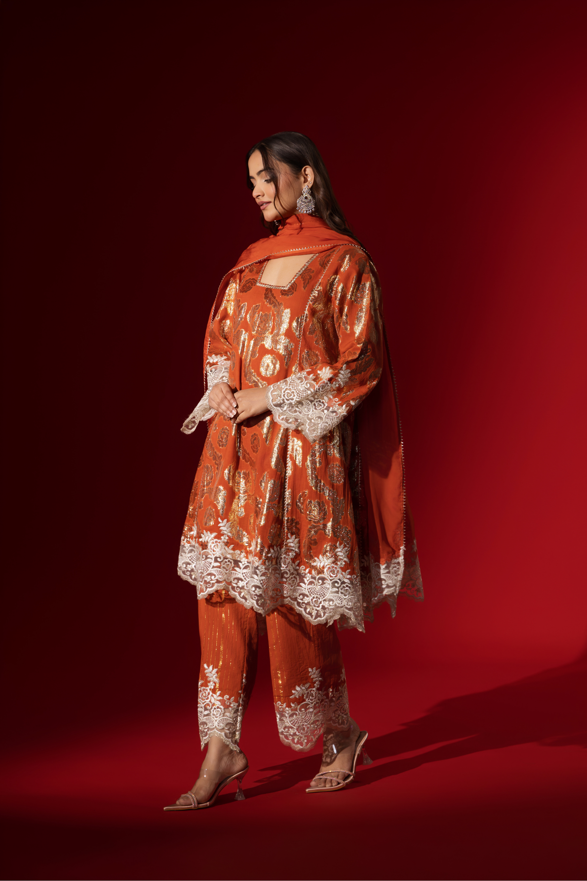 Side view of a woman poses in a orange ethnic shimmer kurta set outfit with intricate white embroidery against a vibrant red backdrop.