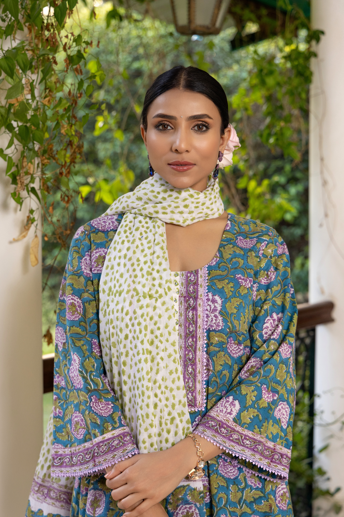 Closeup view of a woman wears a colorful, peacock-printed cotton kurta set with a matching scarf, standing on a checkered floor surrounded by greenery.