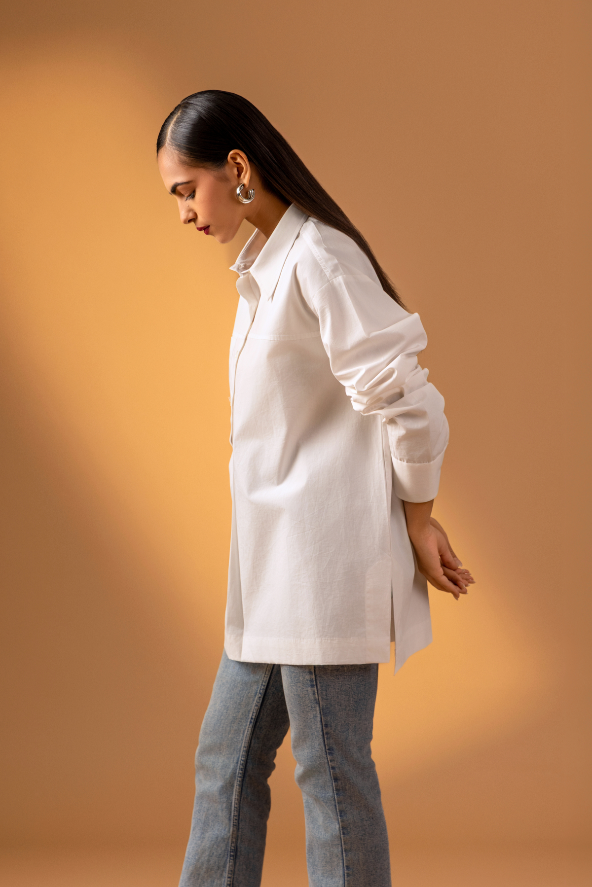 Side view of a woman is dressed in a stylish white button-up shirt with long sleeves, paired with denim jeans, set against a warm beige background.