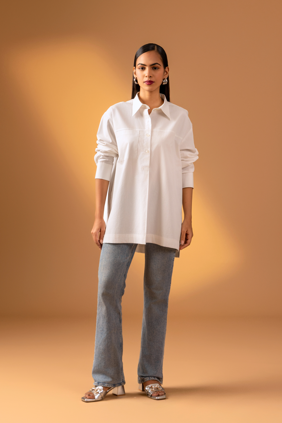 A woman is dressed in a stylish white button-up shirt with long sleeves, paired with denim jeans, set against a warm beige background.