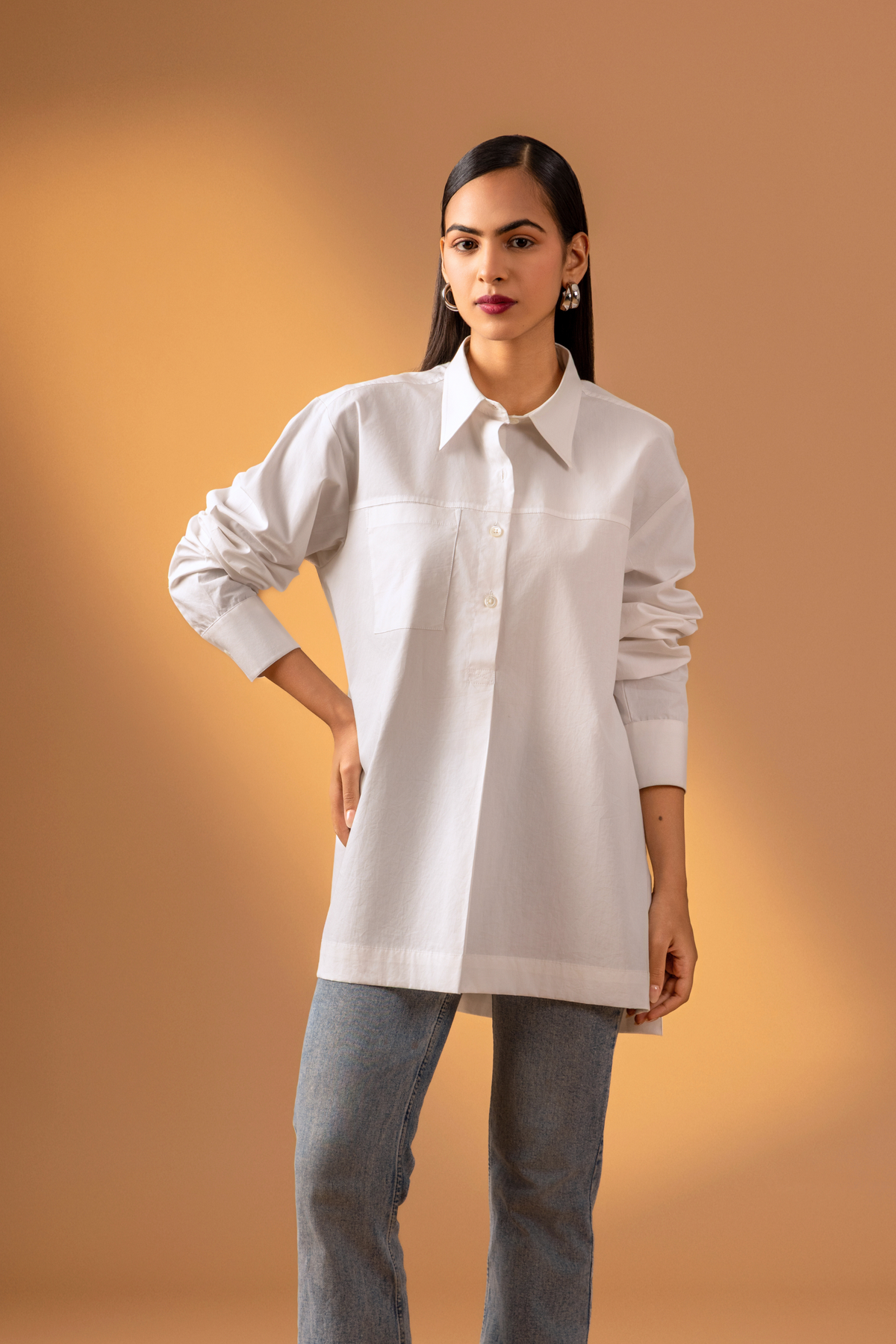 Closeup view of a woman is dressed in a stylish white button-up shirt with long sleeves, paired with denim jeans, set against a warm beige background.