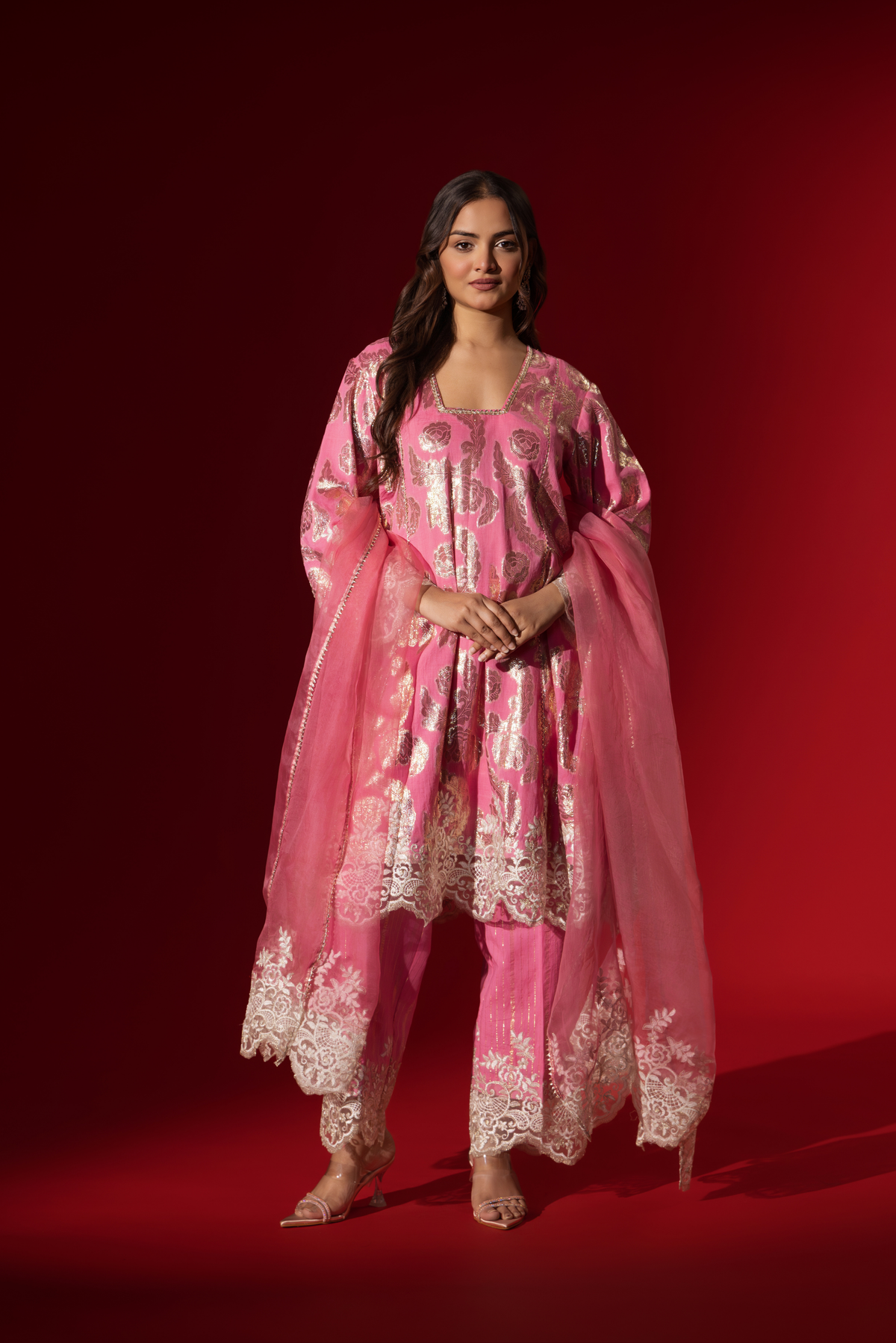 A woman poses in a pink ethnic shimmer kurta set outfit with intricate white embroidery against a vibrant red backdrop.