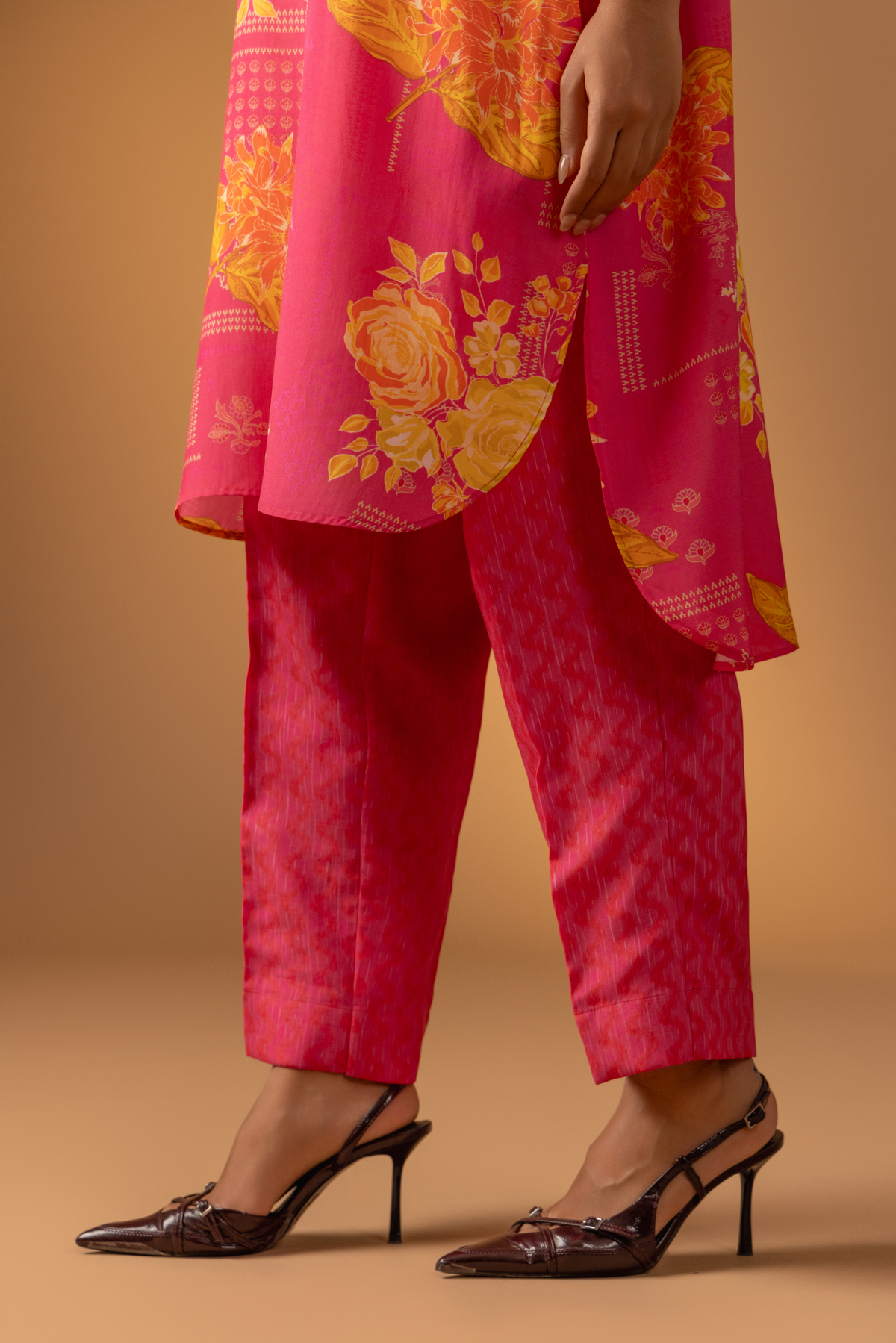 A close-up of a woman wearing a vibrant pink co-ord set with floral patterns and chic brown heels against a soft, warm background.
