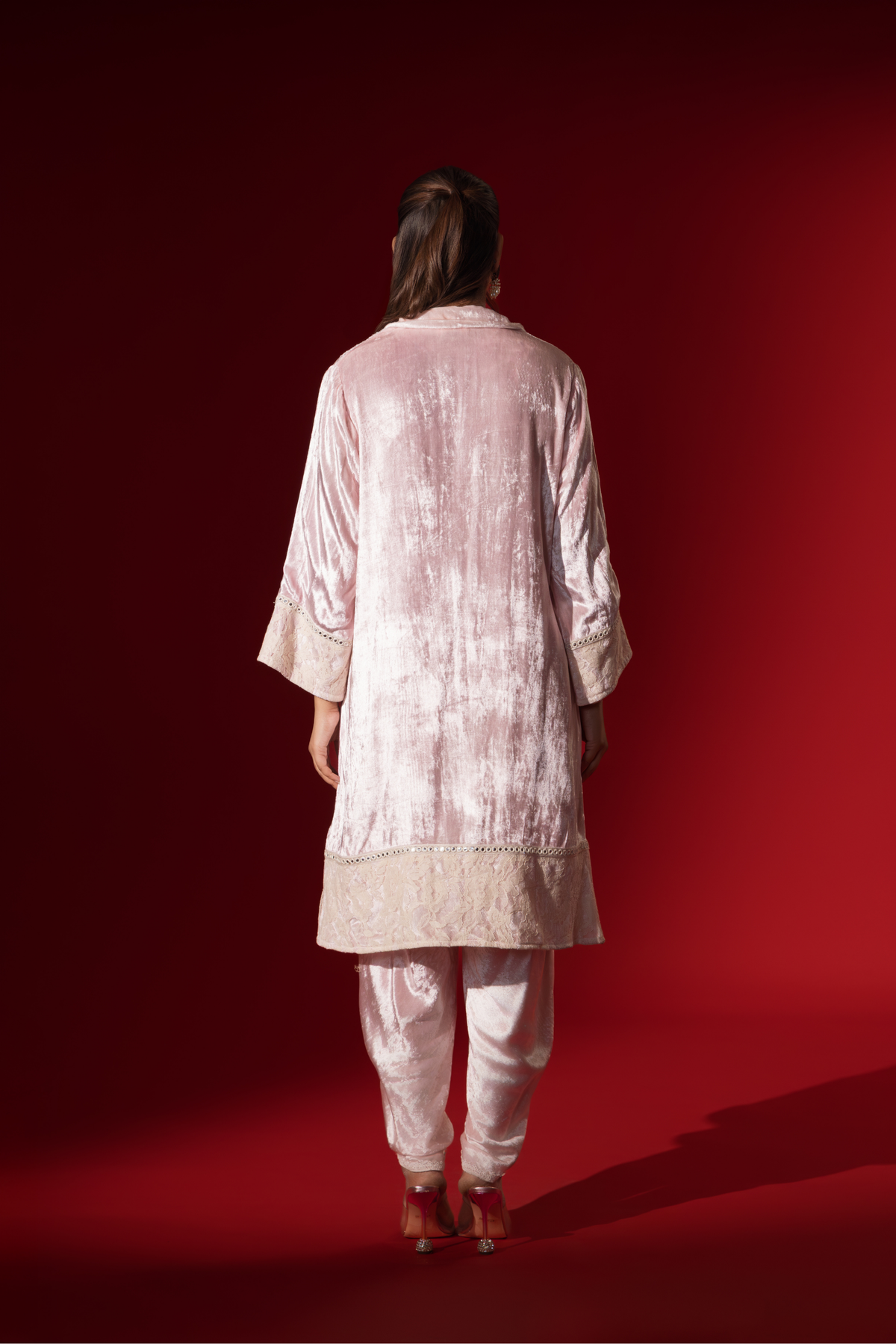 Back view of a woman stands in a champagne pink velvet co-ord set with a long tunic and draped pants, against a vibrant red backdrop.
