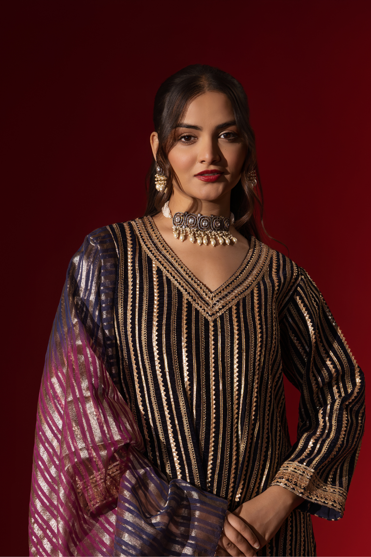 Closeup view of a woman showcases an ethnic plum velvet gota kurta set with golden stripes, paired with a shimmering shawl, against a rich red backdrop.