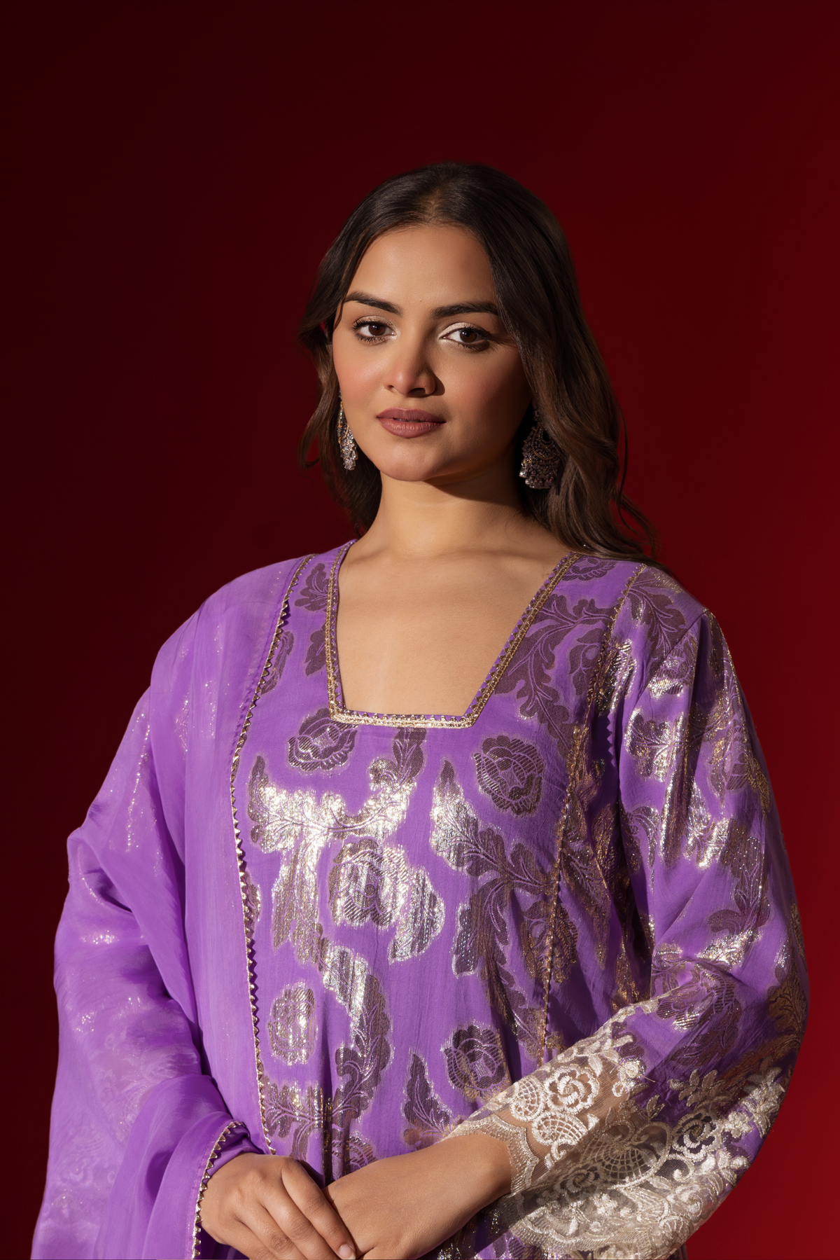 Closeup view of a woman poses in a purple ethnic shimmer kurta set outfit with intricate white embroidery against a vibrant red backdrop.